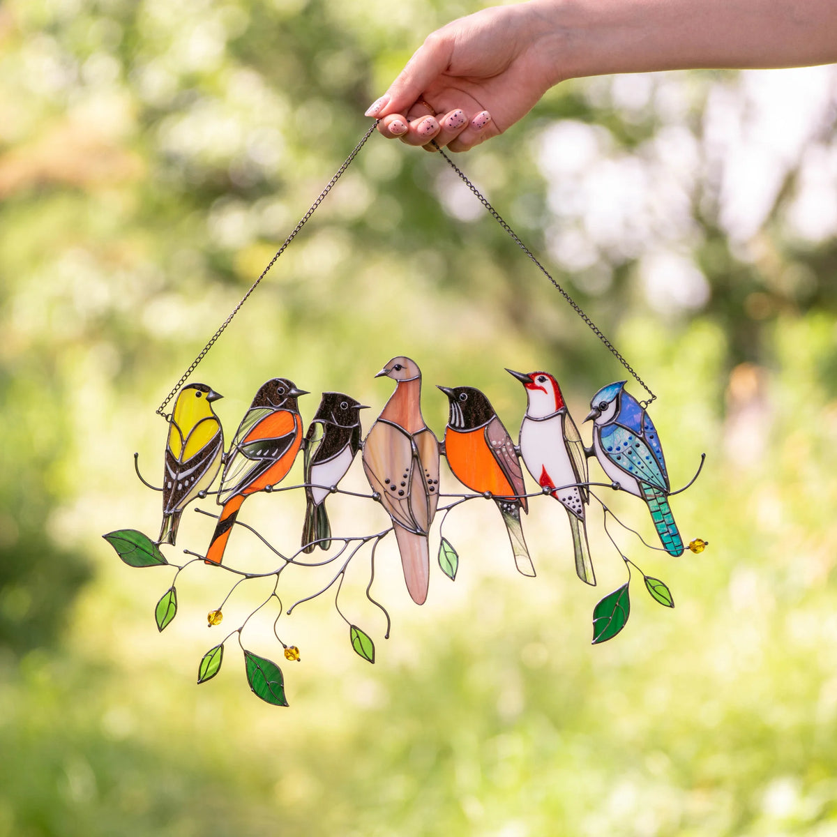 A hand holds a stained glass suncatcher with seven birds on a branch, featuring green leaves and beads, against a blurred garden background.