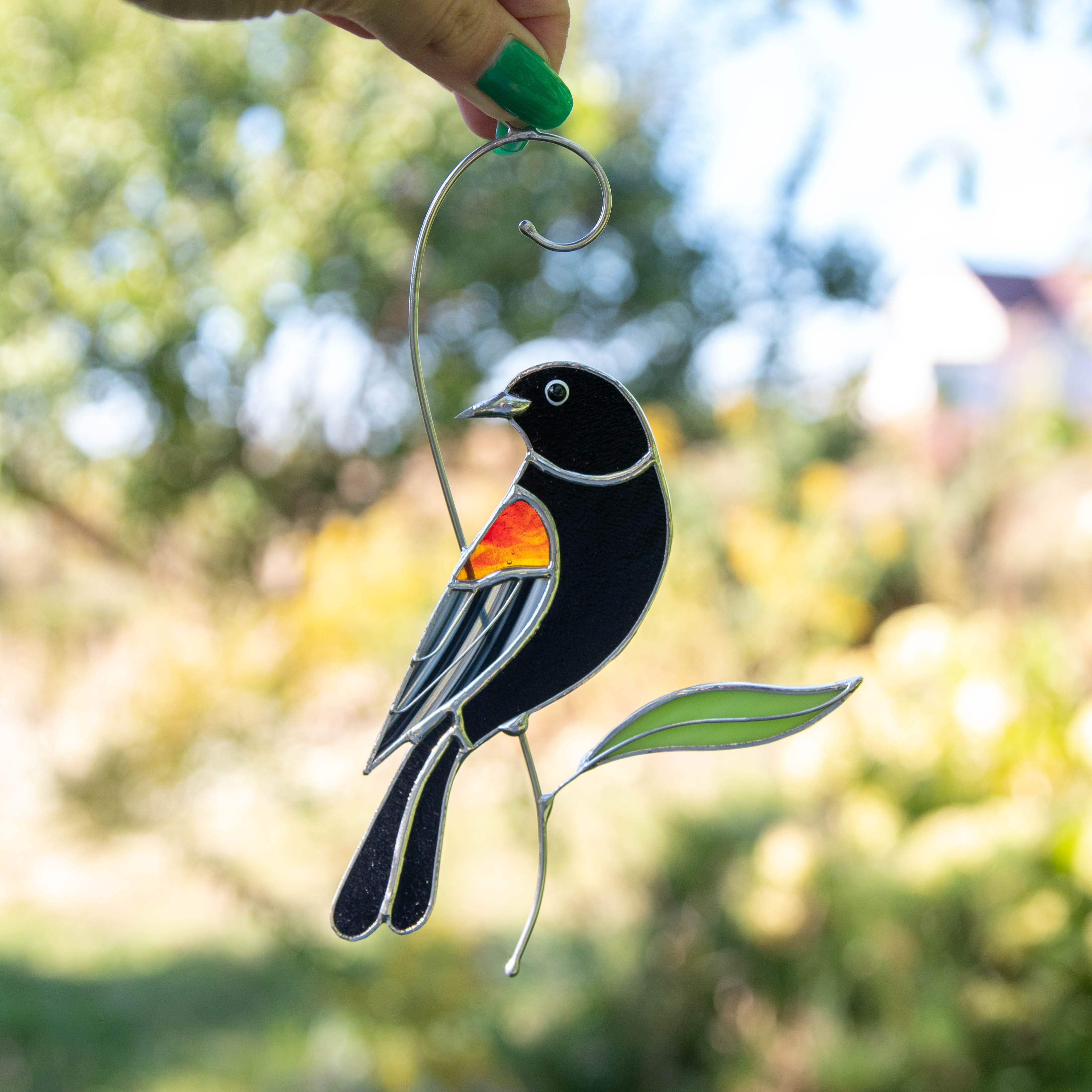 Stained glass blackbird with orange wing and green leaf hanging outdoors.