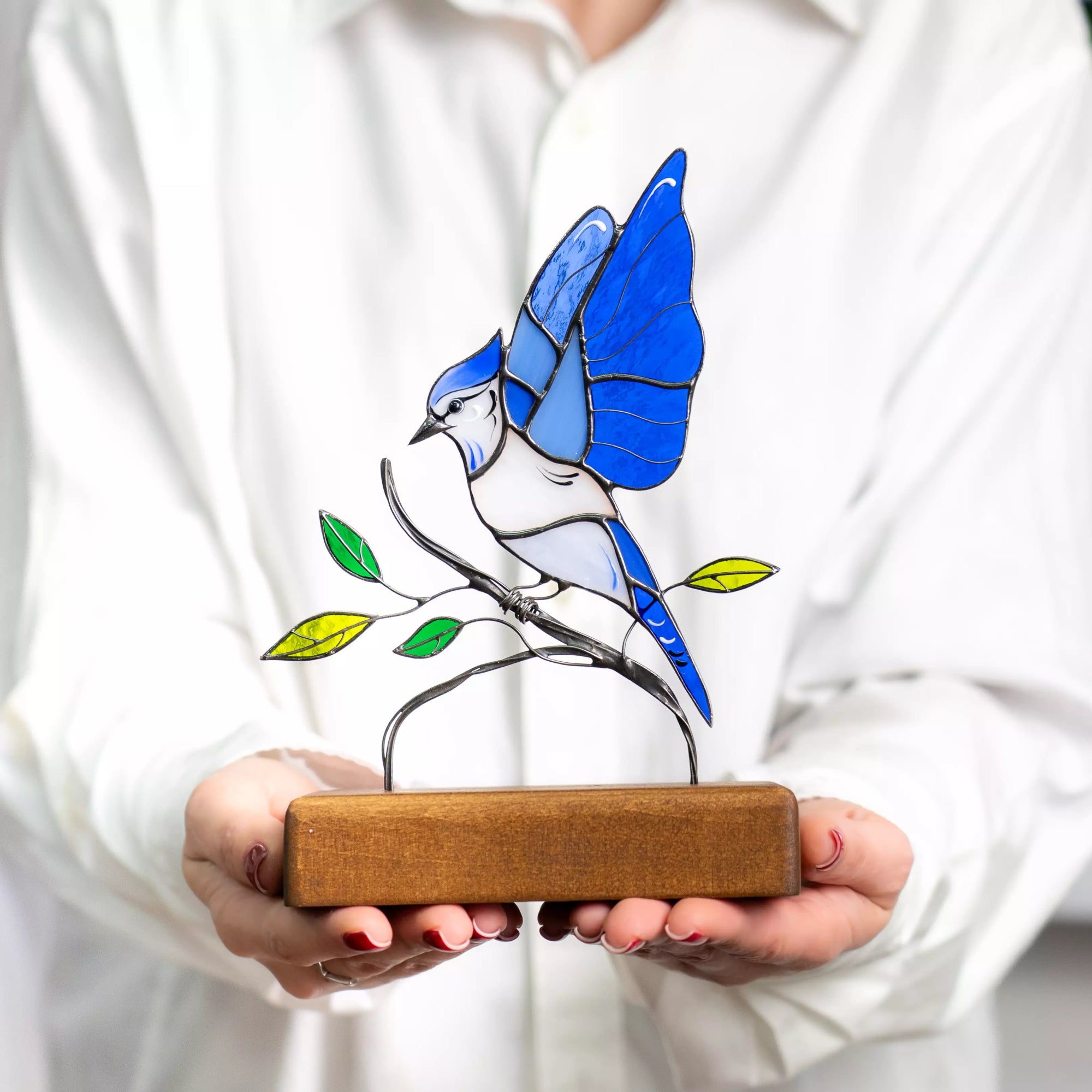Woman in white shirt holding stained glass blue jay figure with raised wings and leafy branch on wood stand