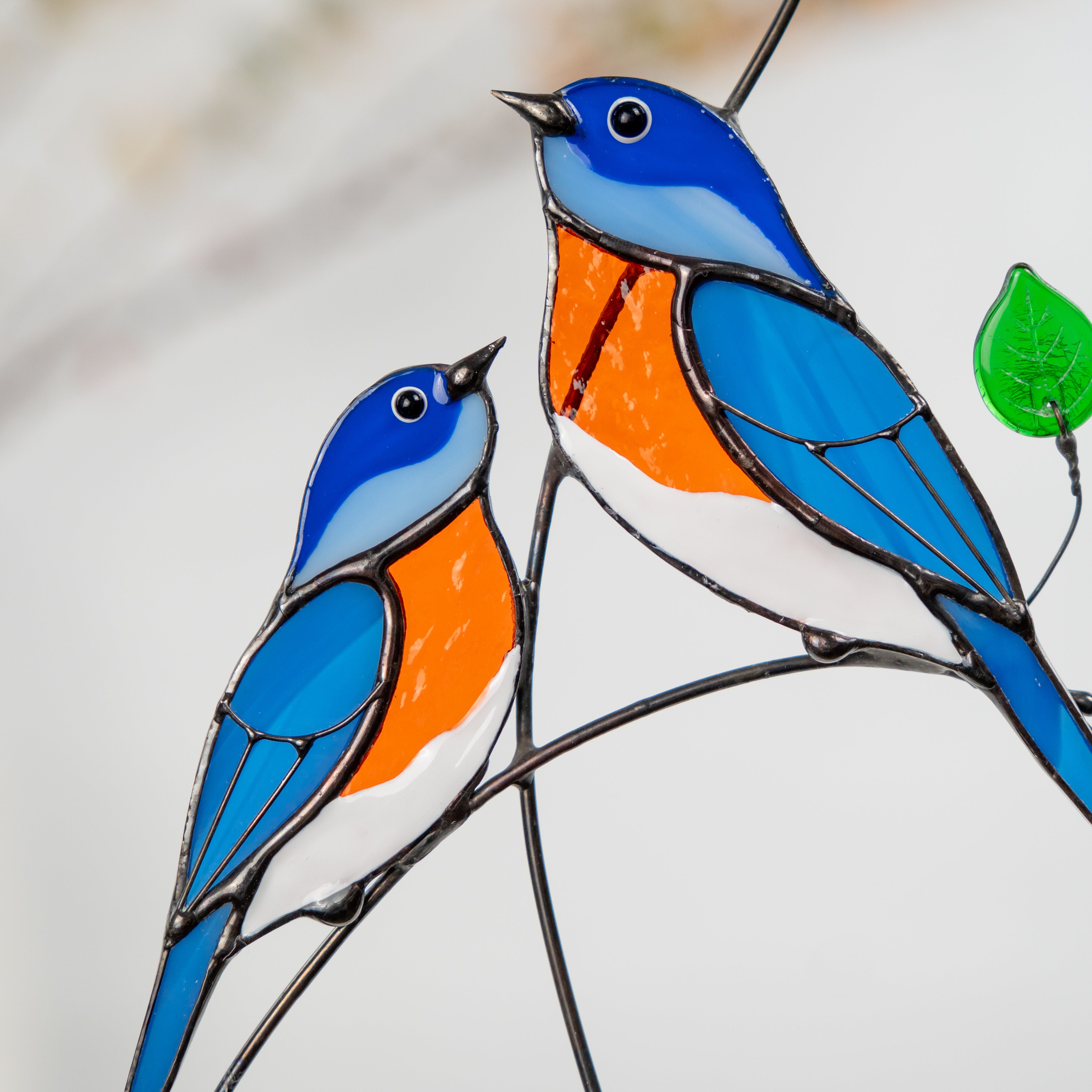 Close-up of two stained glass bluebirds in blue and orange glass perched on a branch with green leaf