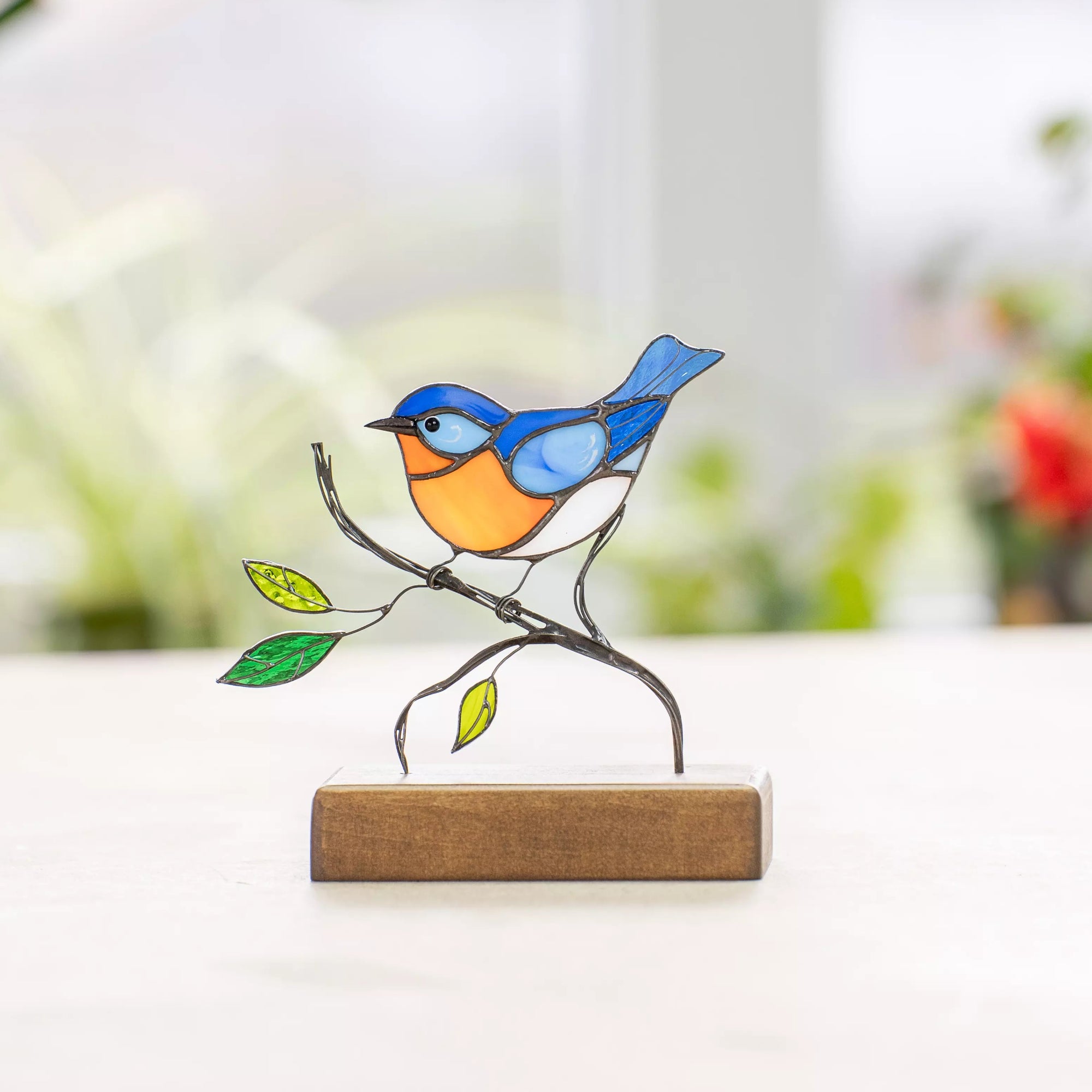 Colorful stained glass bluebird with orange belly and blue wings standing on a curved wire branch with green leaves and wooden base.