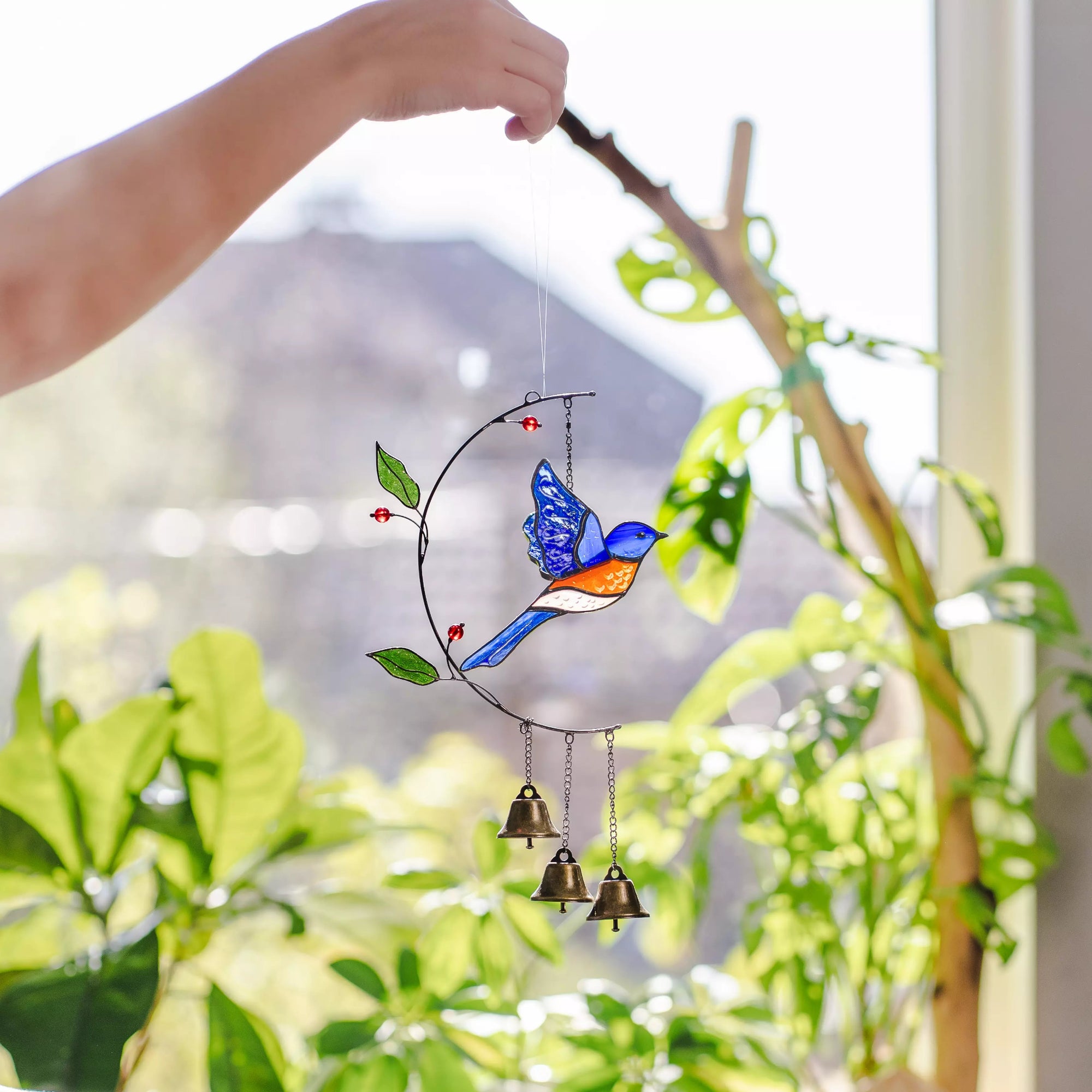 Stained glass bluebird suncatcher with bells hanging from a branch with green leaves.