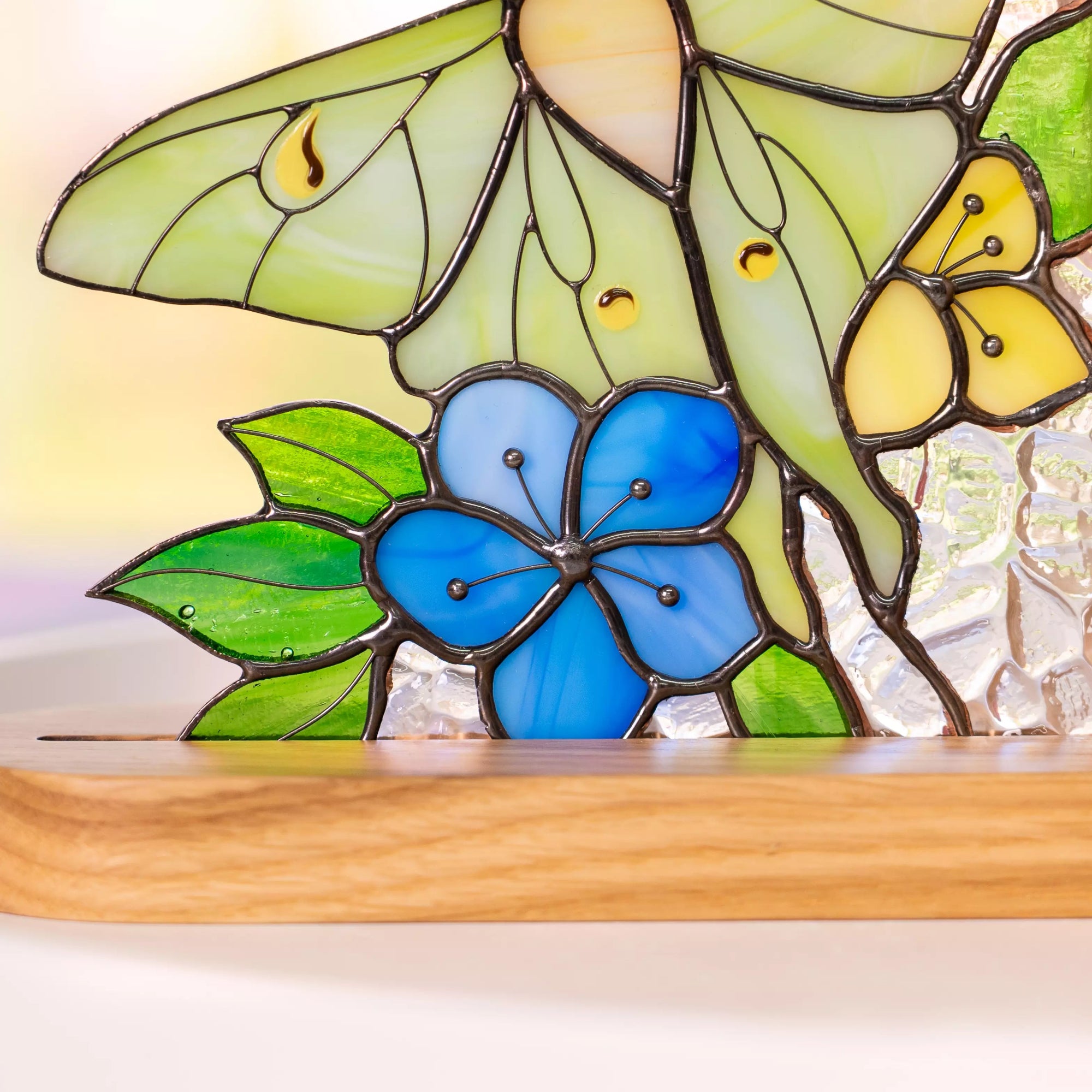 Close-up of stained glass luna moth bookend with blue flower, green leaves, and textured clear glass