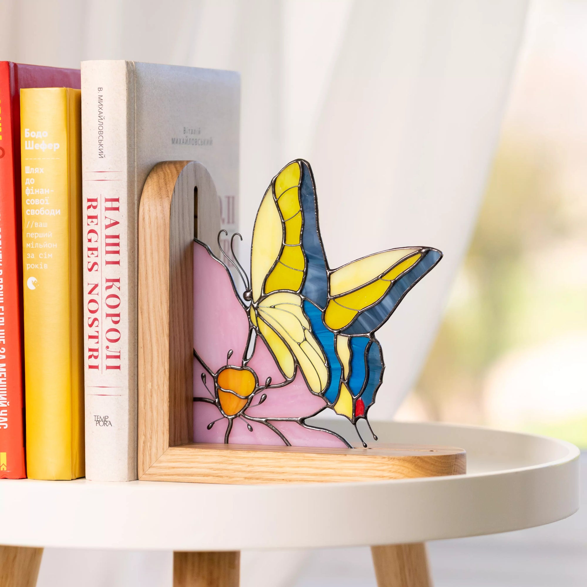 Stained glass butterfly bookend displayed in a wooden stand beside books, featuring yellow and blue wings with a pink glass base, shown as decorative shelf home decor.