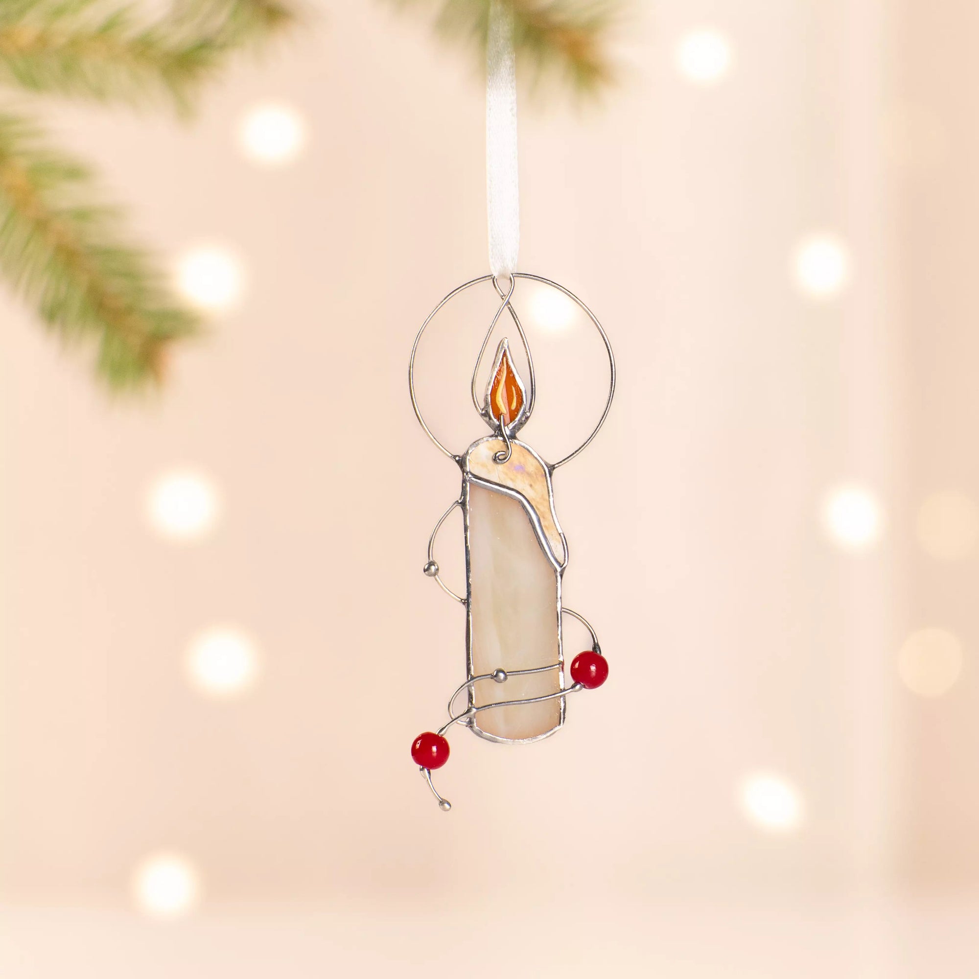 Stained glass candle ornament hanging near pine branch with glowing background lights.