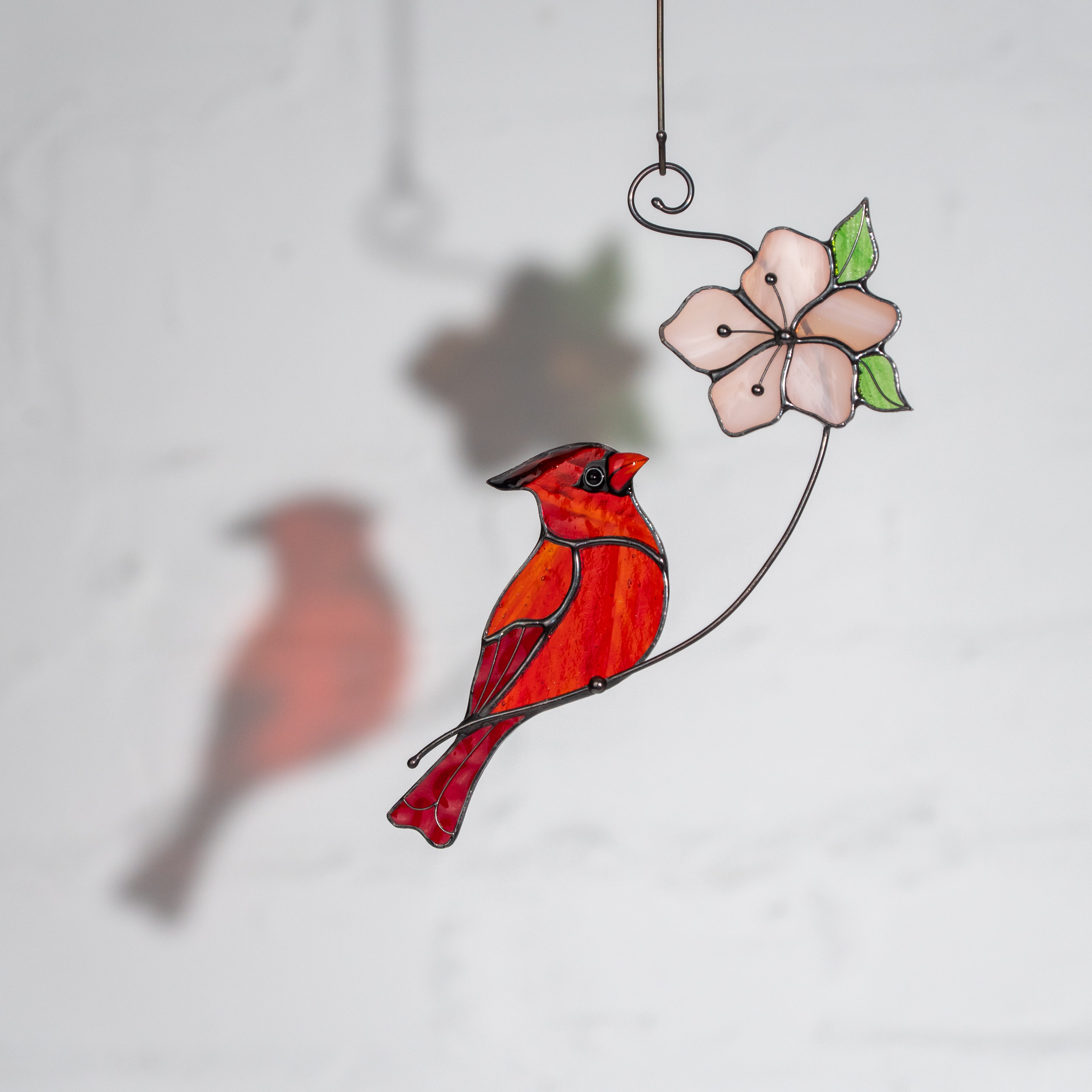 Bright red stained glass cardinal perched beside a soft pink flower with green leaves, casting a delicate shadow on the wall.
