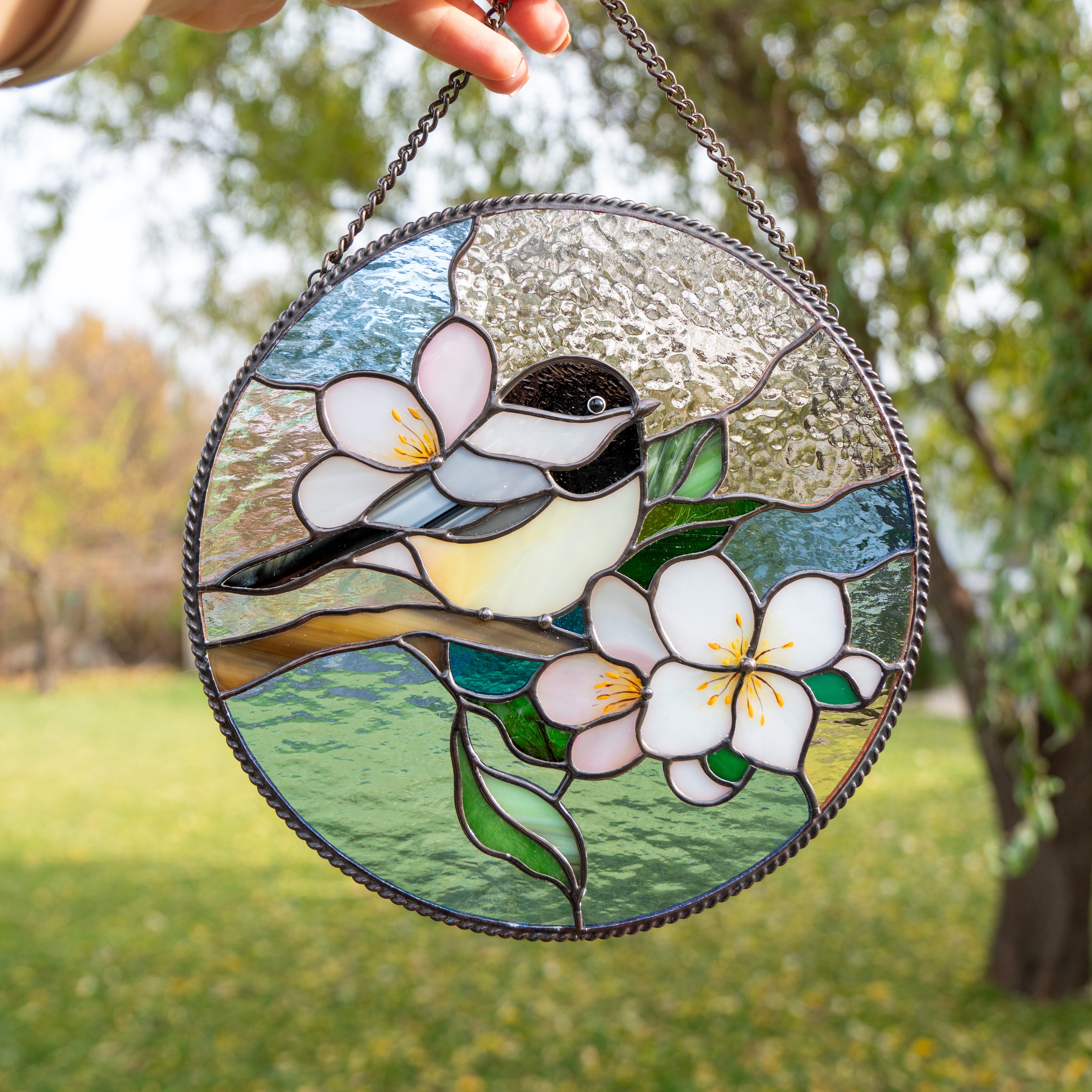 A round stained glass panel featuring a black-capped chickadee perched on a branch with three pink and white flowers against a pastel sky