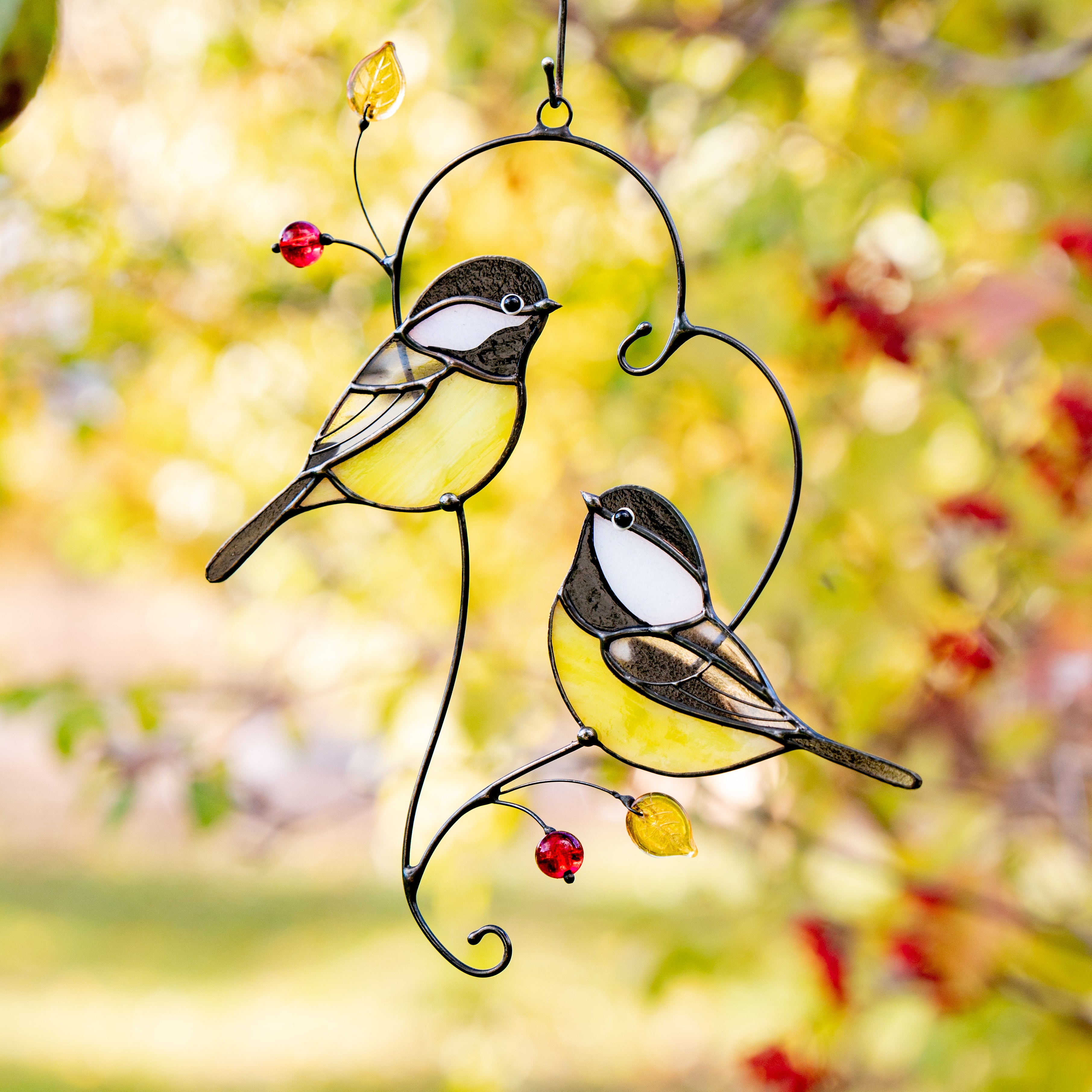 Two stained glass chickadees with yellow bellies and red beads, hanging among autumn leaves in soft golden light