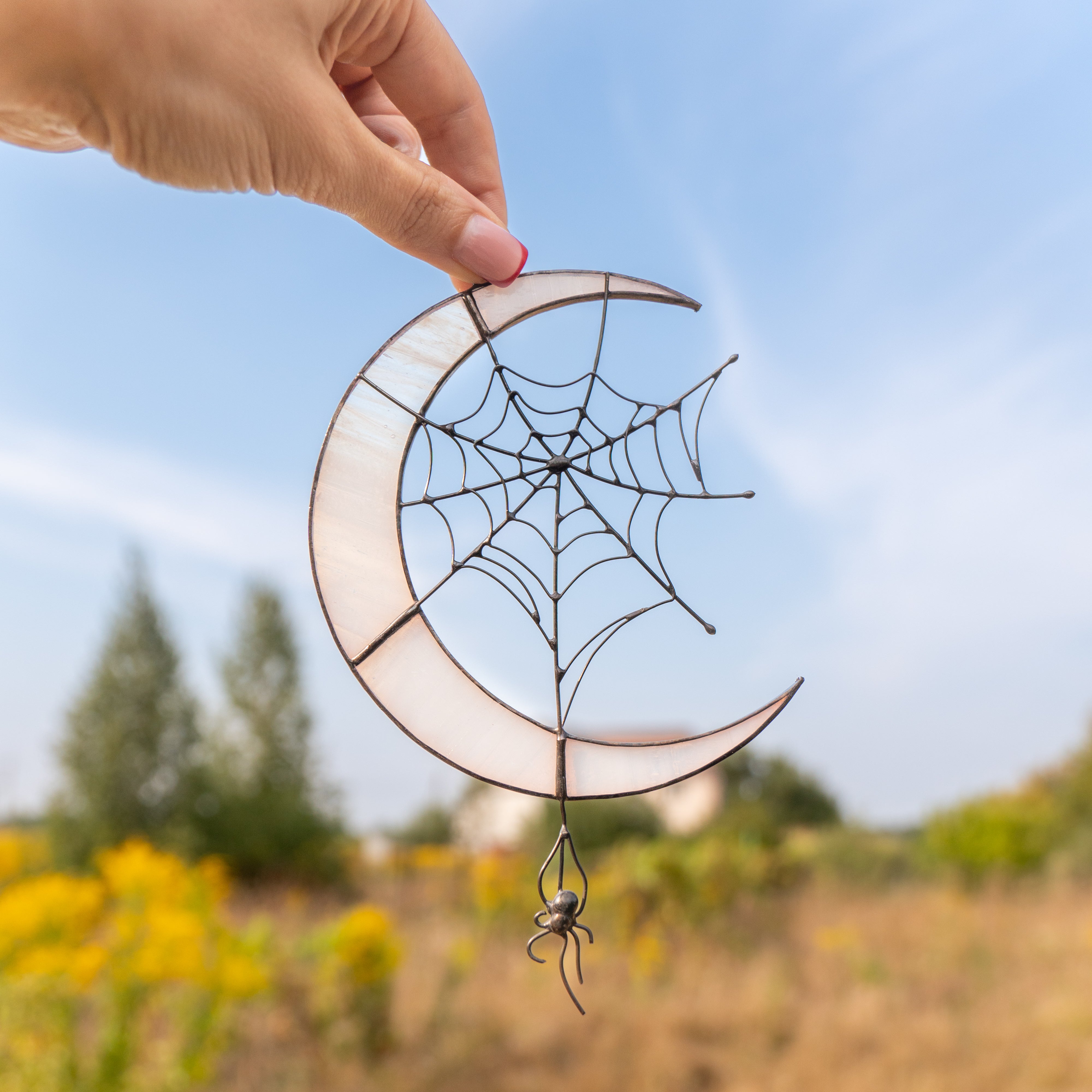 Spider web stained glass crescent moon held against a blue sky and yellow wildflowers.