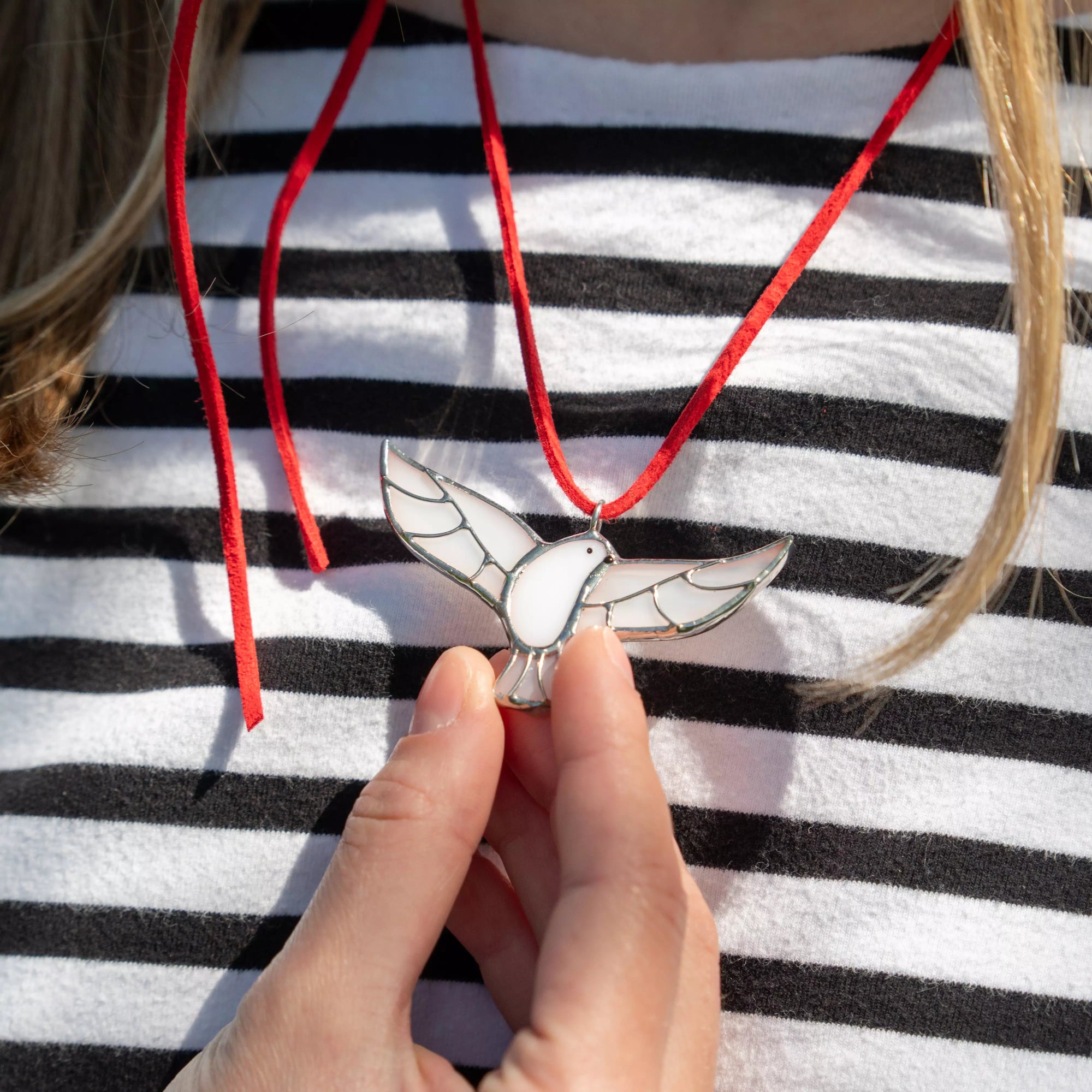 Close-up of a person holding a stained glass dove pendant on a red ribbon worn around the neck over a striped shirt