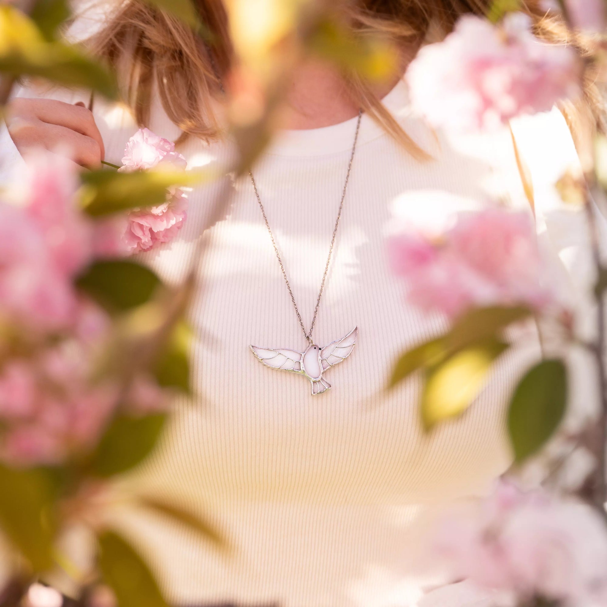 Woman wearing white top and stained glass dove pendant necklace among blooming pink flowers