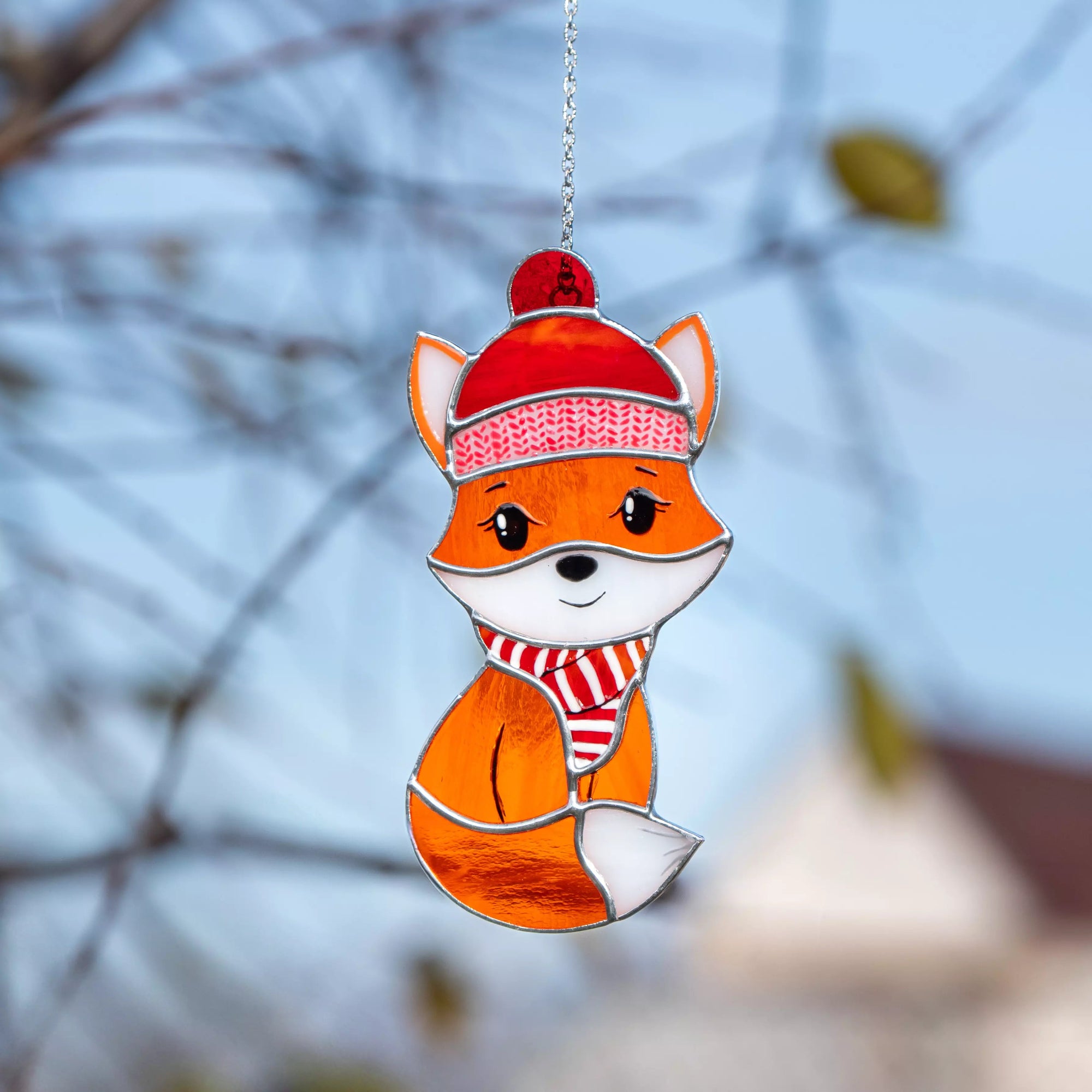 Stained glass fox suncatcher wearing a red hat and scarf against a blurred natural background