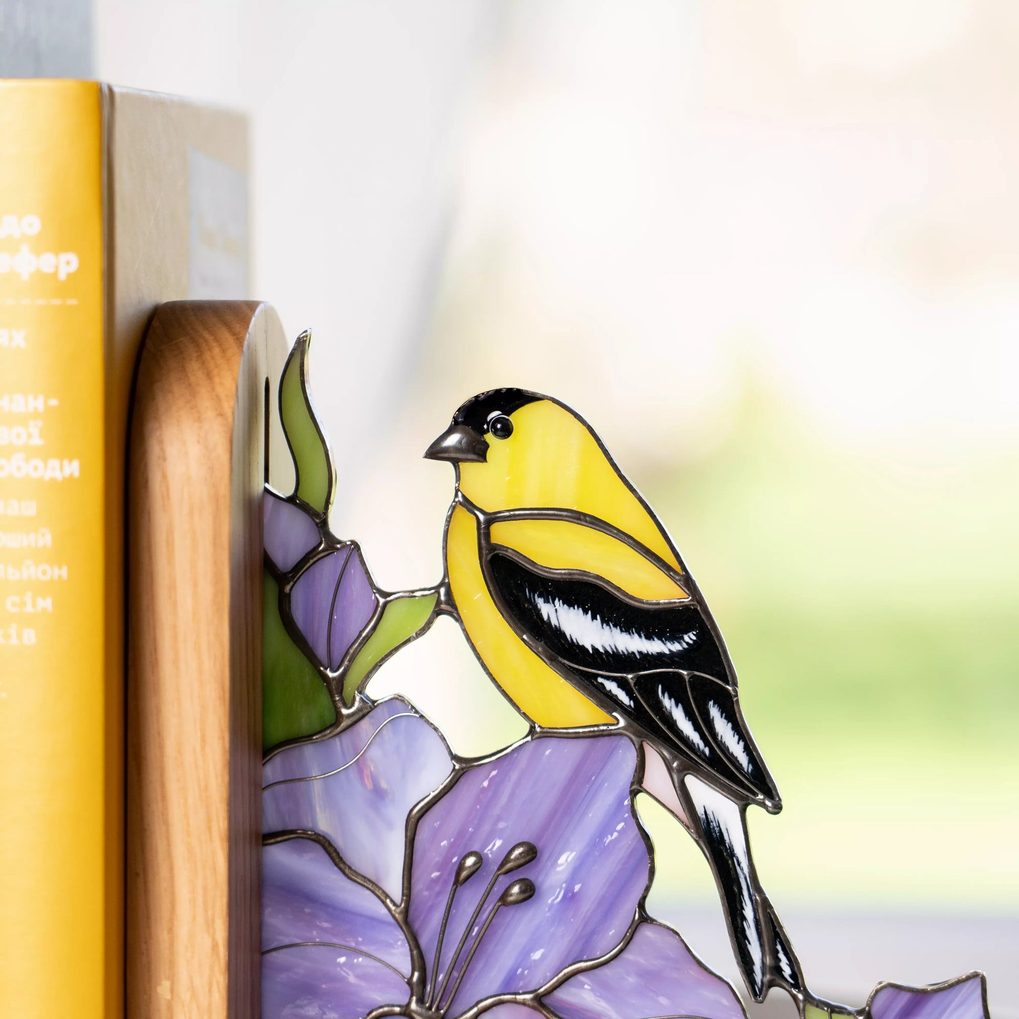 Stained glass goldfinch bird with bright yellow and black feathers perched on a purple flower, handcrafted glass decor displayed as a bookend on a wooden shelf near books and a window