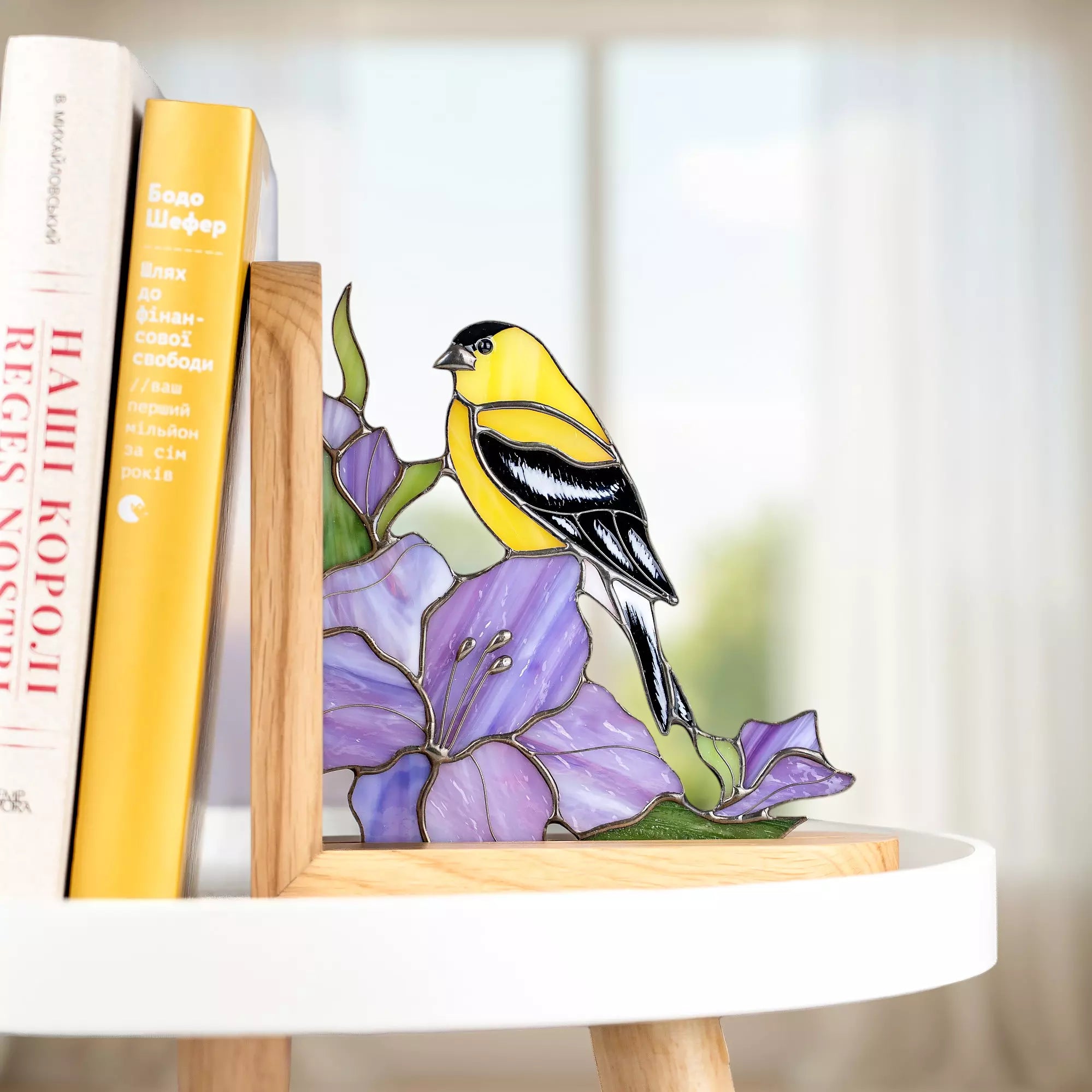 Handcrafted stained glass goldfinch with expressive yellow and black coloring, resting on a purple flower, used as a decorative bookend that adds artistic charm to shelves and reading spaces