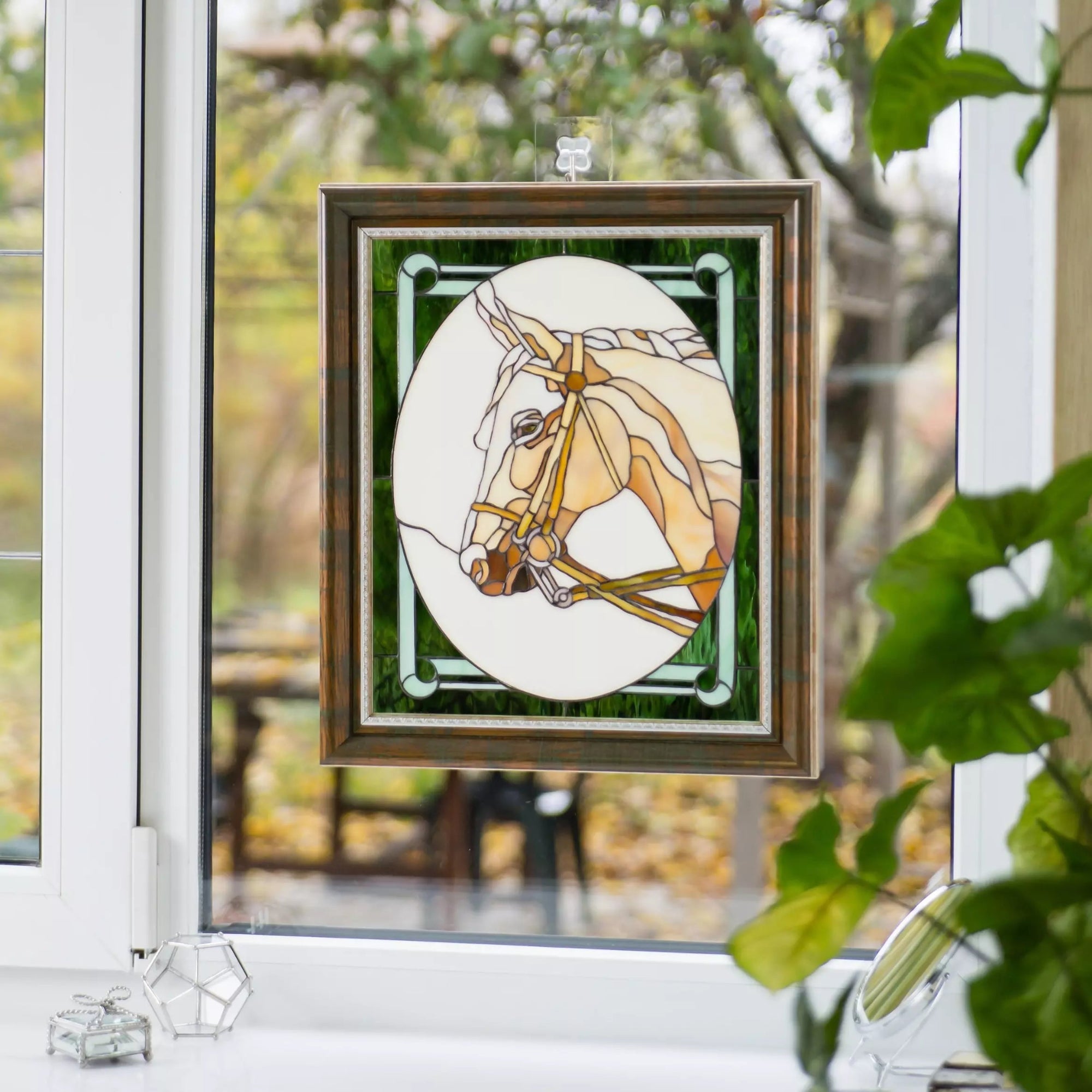 Horse stained glass panel suspended on window with sunlight shining through and greenery outside.
