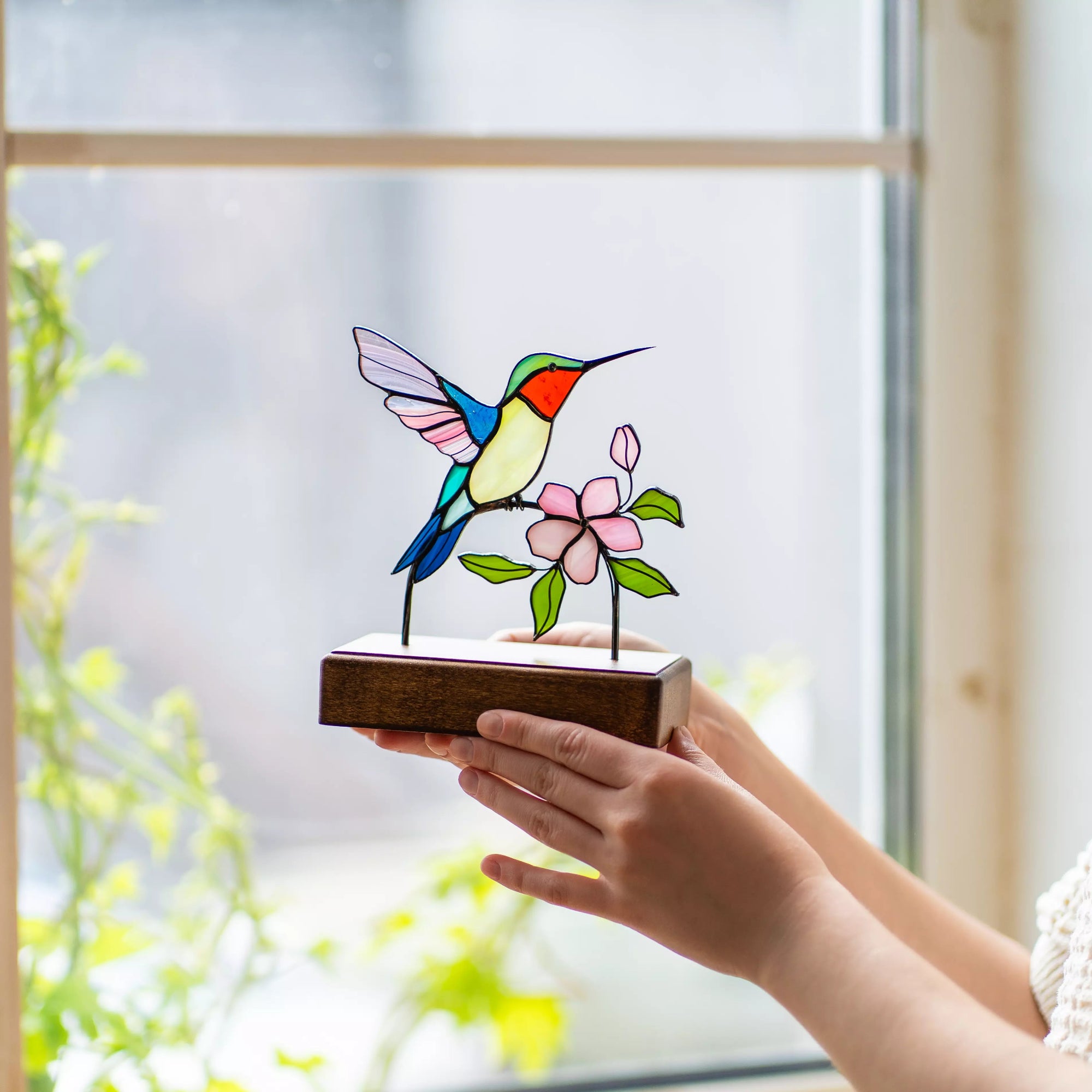 Stained glass hummingbird with sakura flower decoration held by a hand with a window background