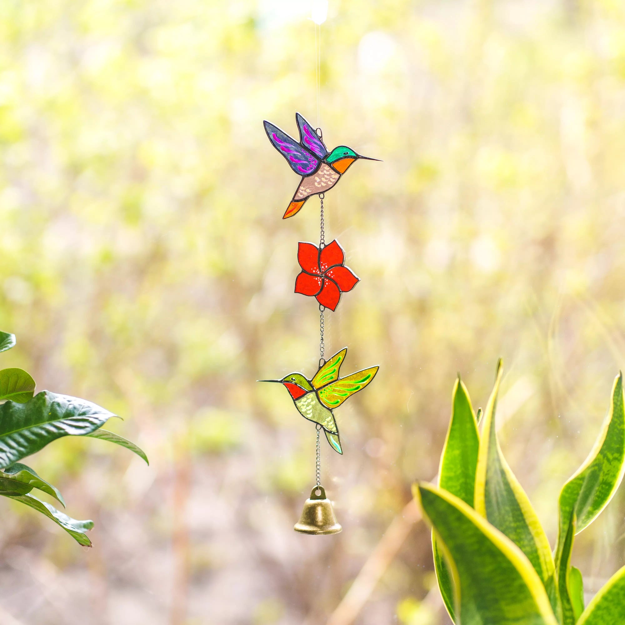 Colorful stained glass hummingbird and flower hanging decoration with a bell against a blurred green outdoor background.