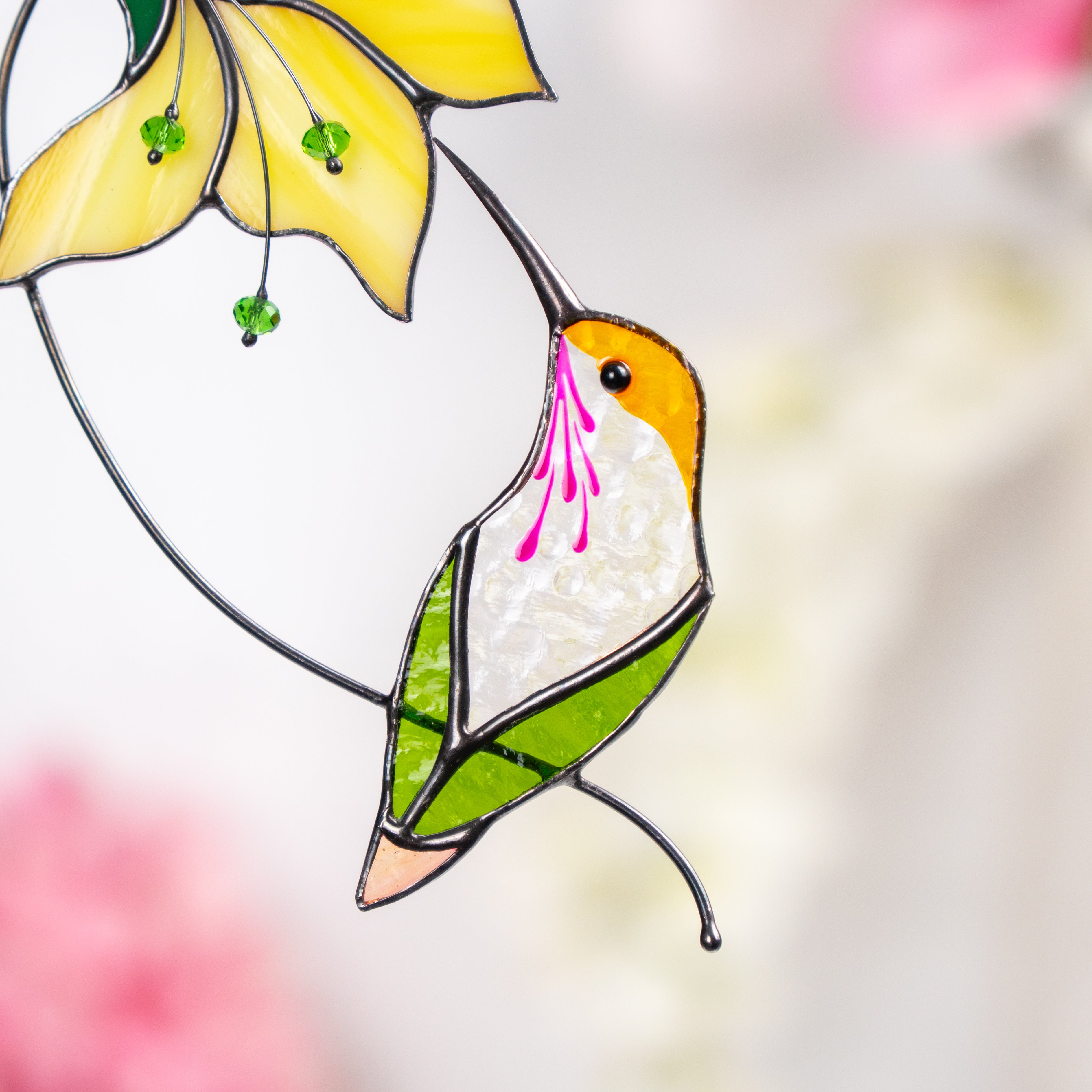 Close-up of a stained glass hummingbird with white, green, and amber tones sipping from a yellow flower.