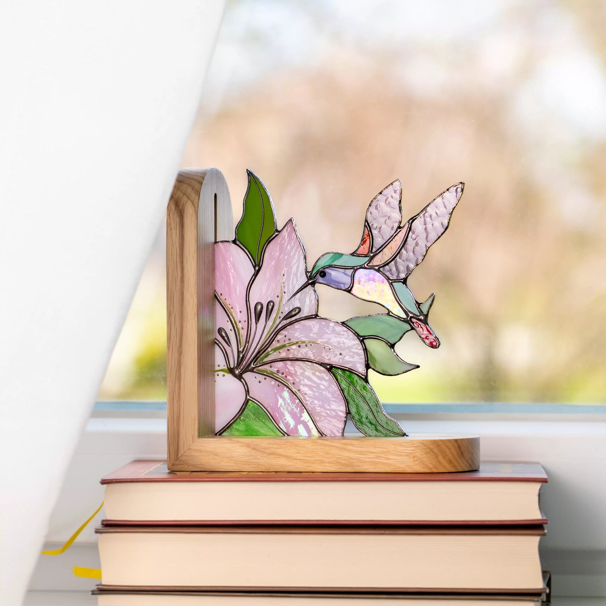 Stained glass hummingbird and pink flower bookend placed on stacked books by window
