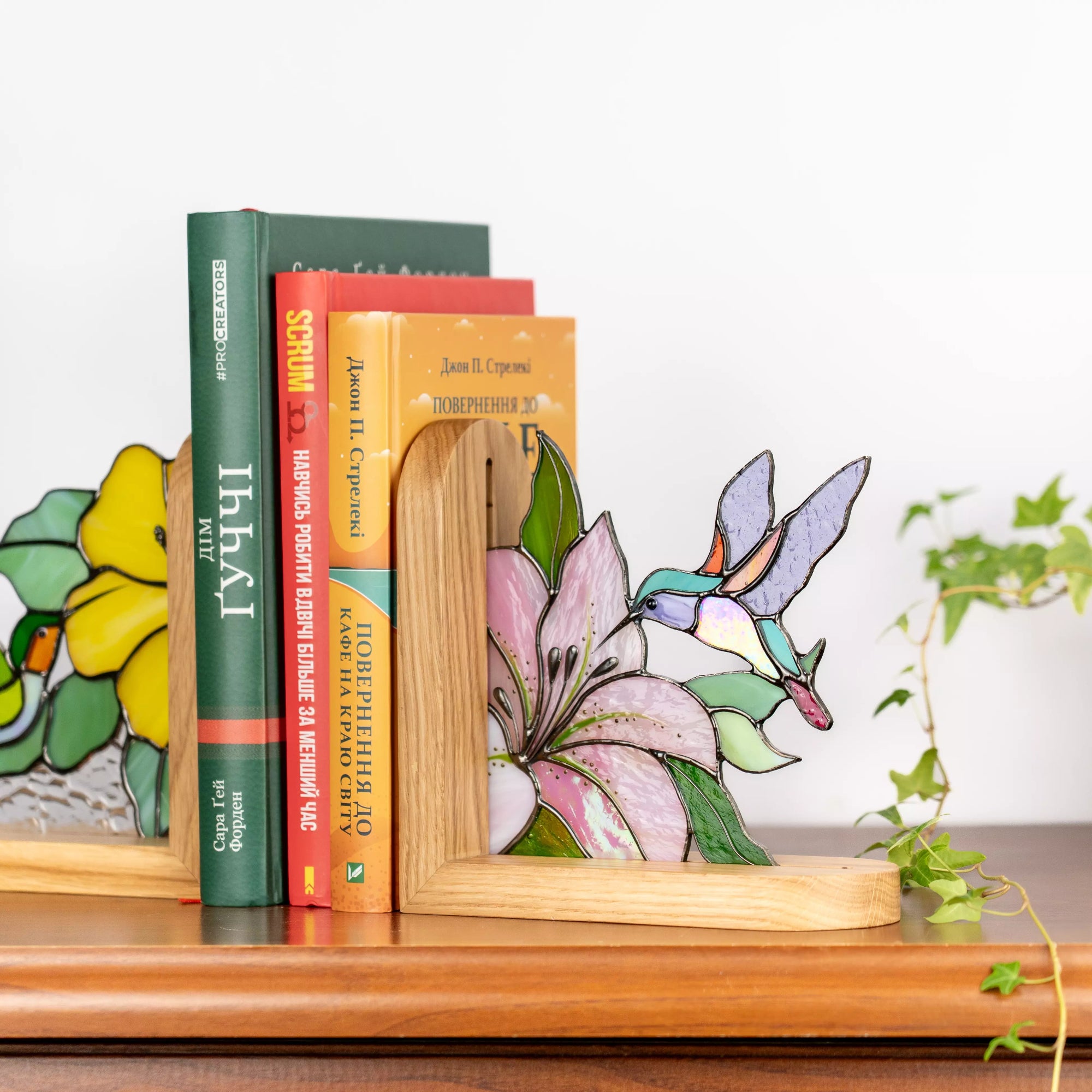 Stained glass hummingbird and pink flower bookend beside books with trailing green plant