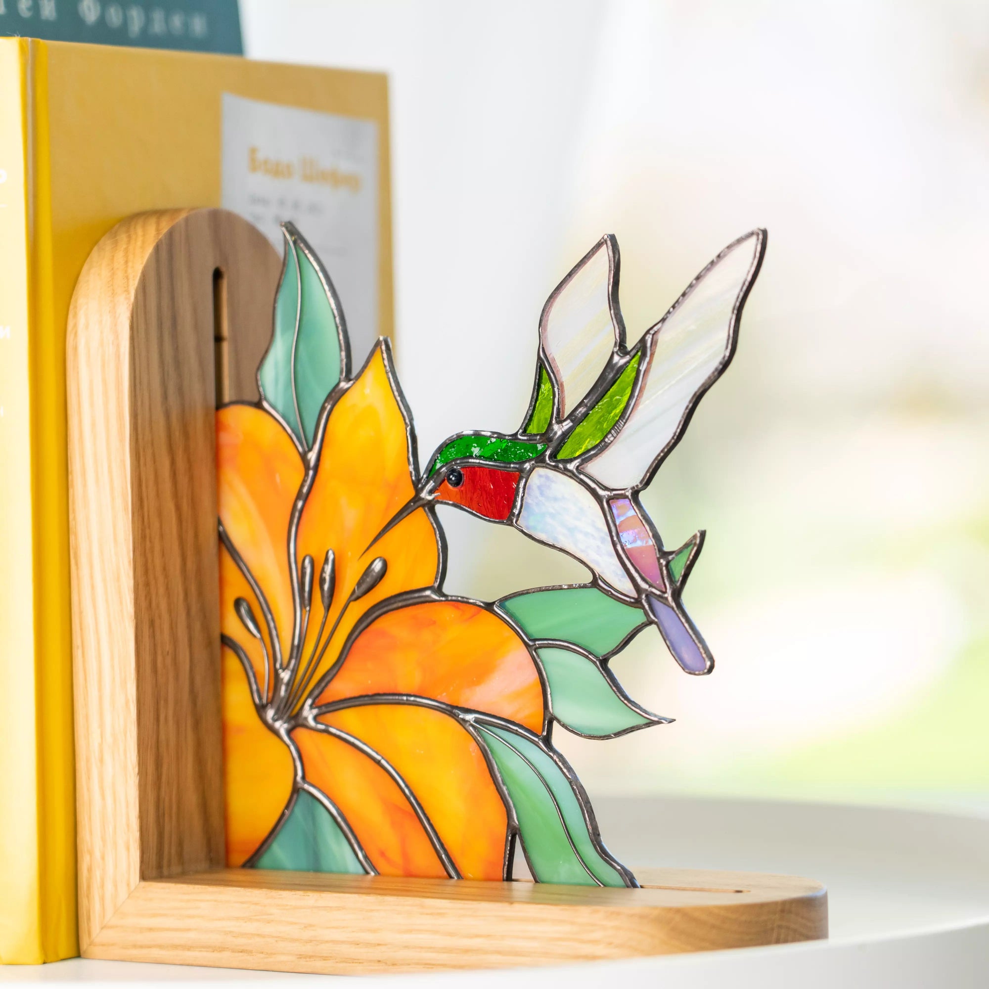Stained glass hummingbird bookend with orange flower and green leaves beside yellow books