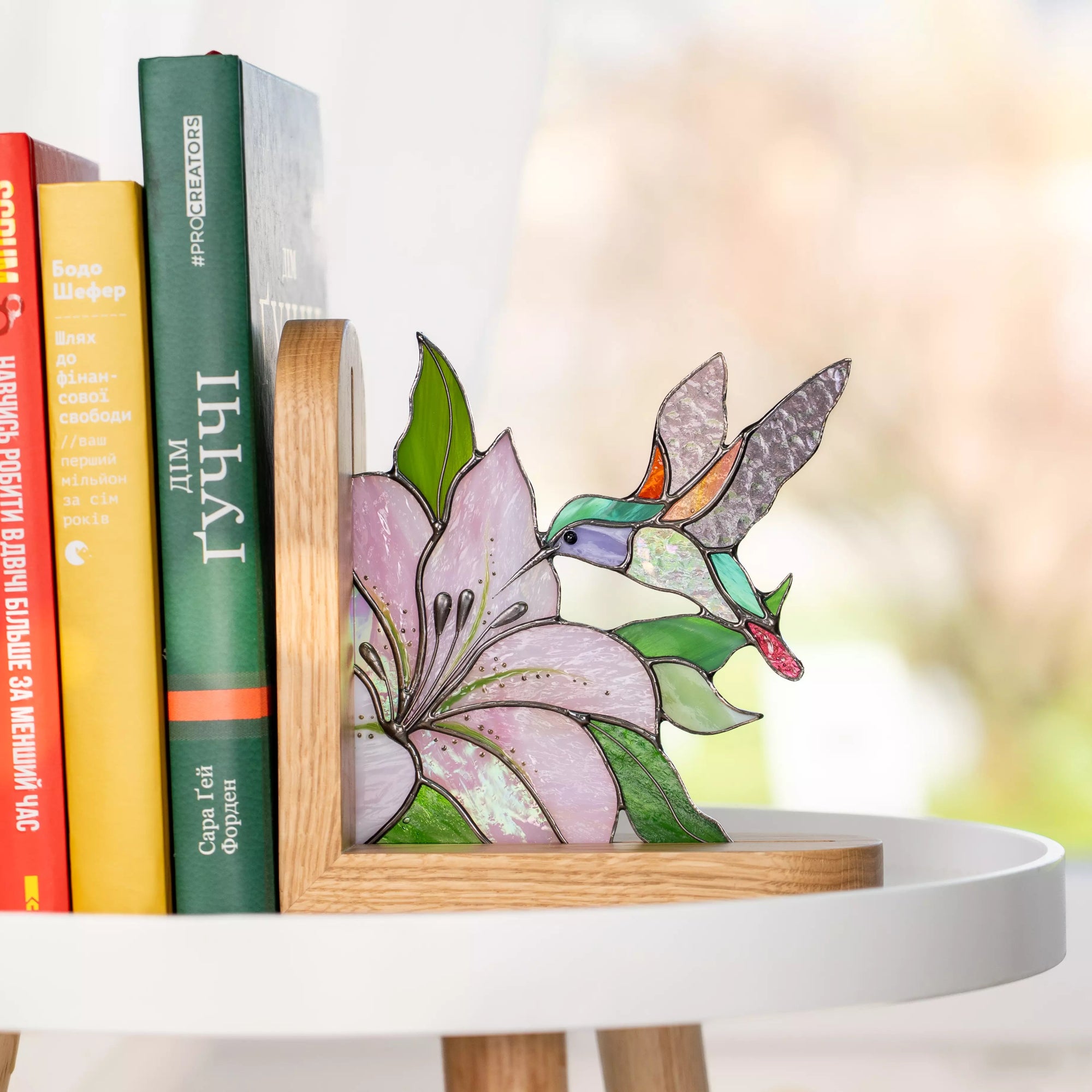 Stained glass hummingbird flying toward a pink flower bookend displayed next to colorful books