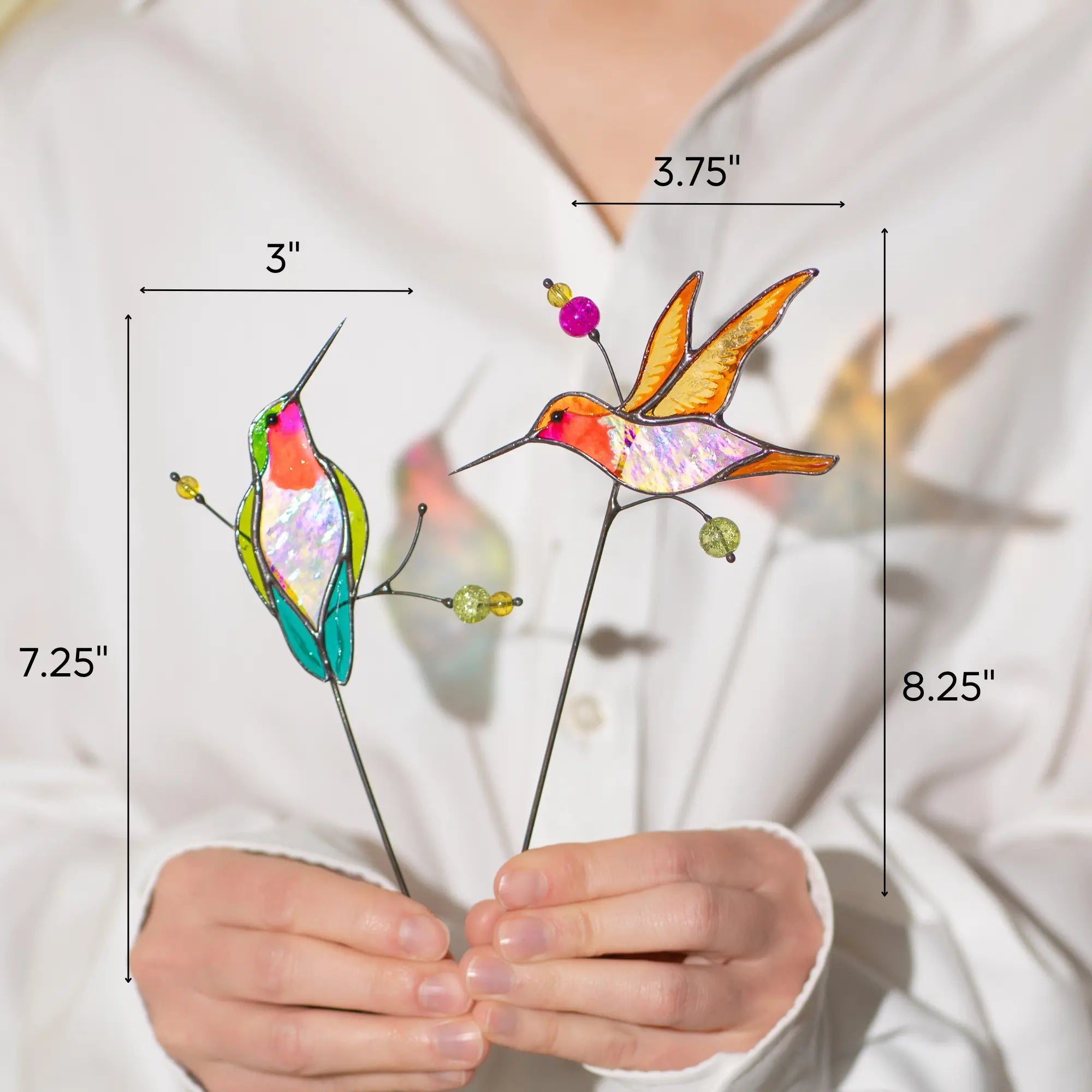 Stained glass hummingbird plant stakes held by a person with measurements indicated: 3 x 7.25 and 3.75 x 8.25 inches
