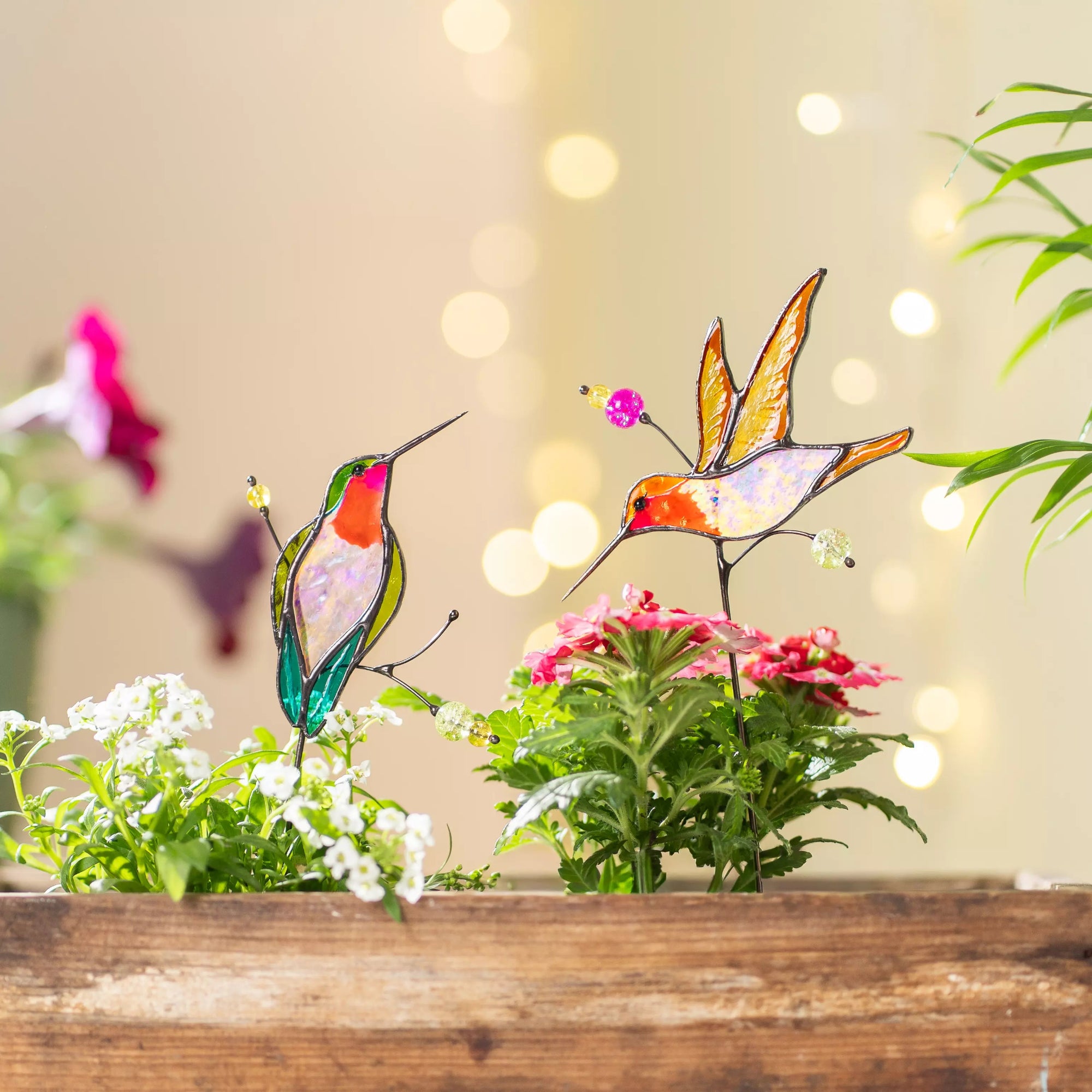 Stained glass hummingbird plant stakes on a wooden surface with flowers and blurred lights in the background