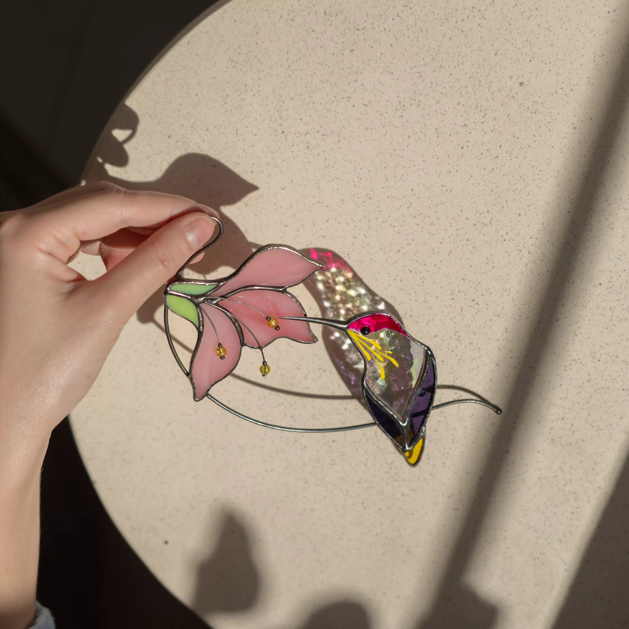 Hand holding a stained glass hummingbird with purple wings and pink flower on a beige surface, casting a soft shadow