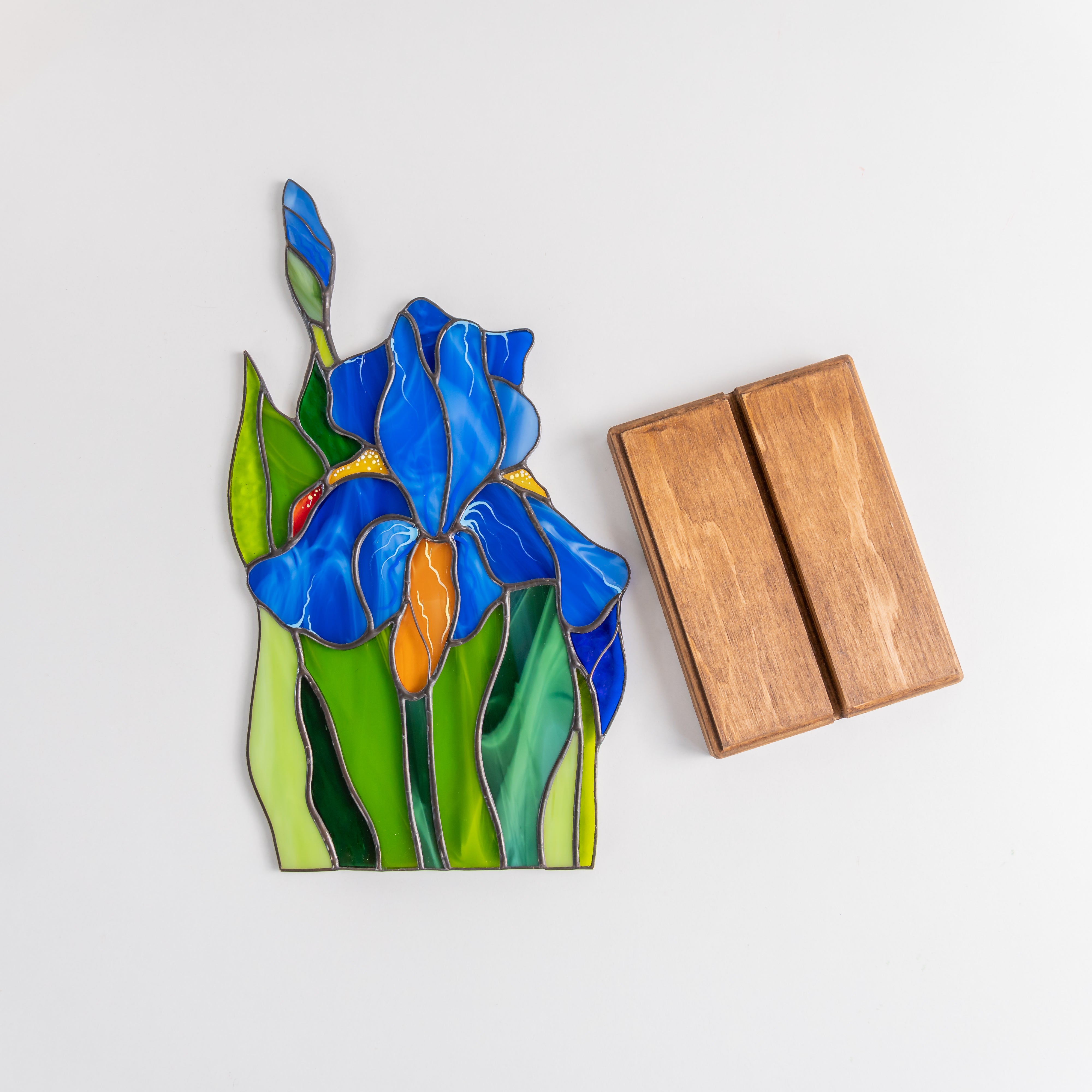 Iris flower stained glass panel laid flat beside two-piece wooden display stand on white surface.