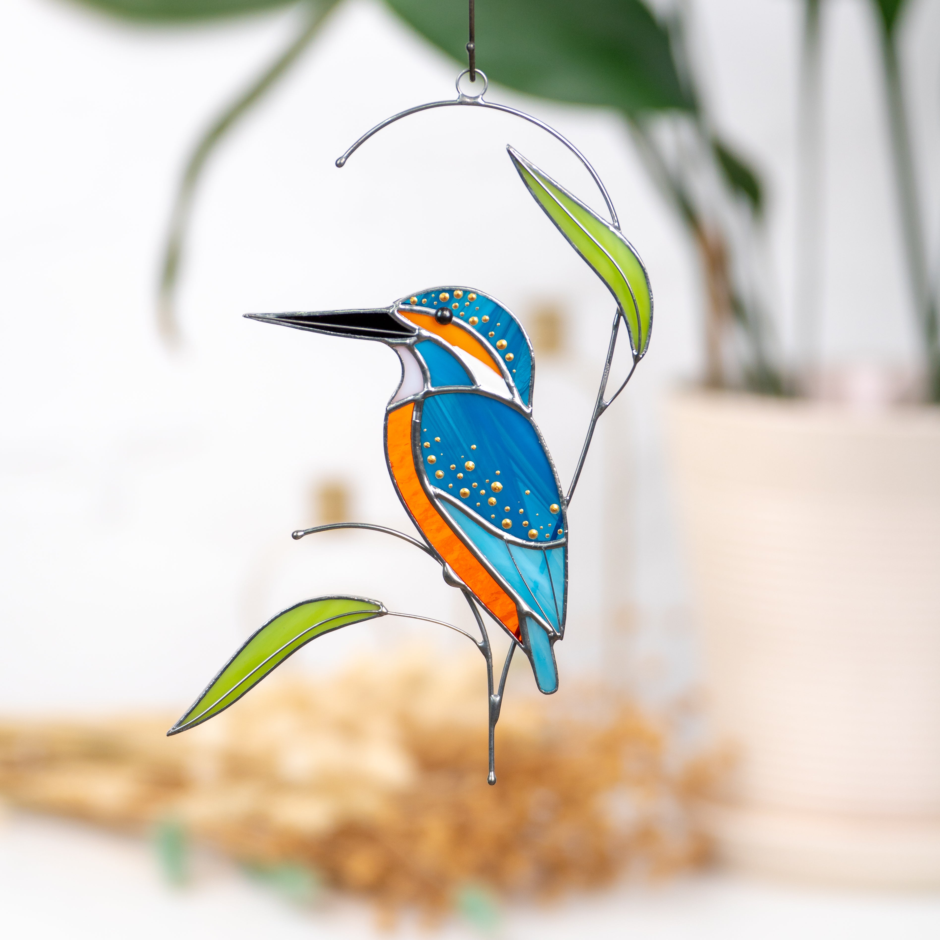 Colorful stained glass kingfisher perched on a metal branch with green leaves, hanging indoors near blurred decor.