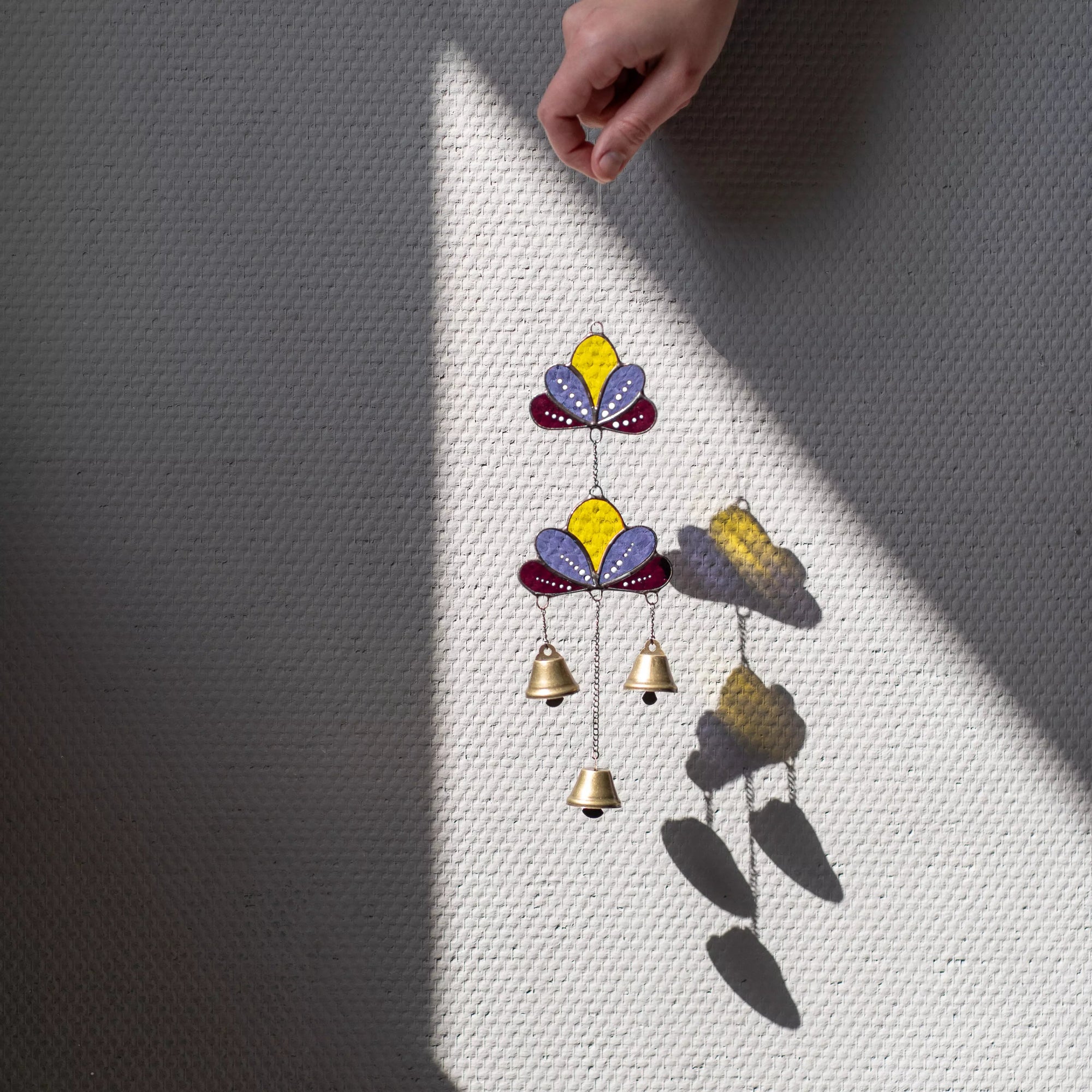 A person's hand holds a stained glass lotus wind chime against a neutral wall, creating colorful shadows in the direct sunlight.