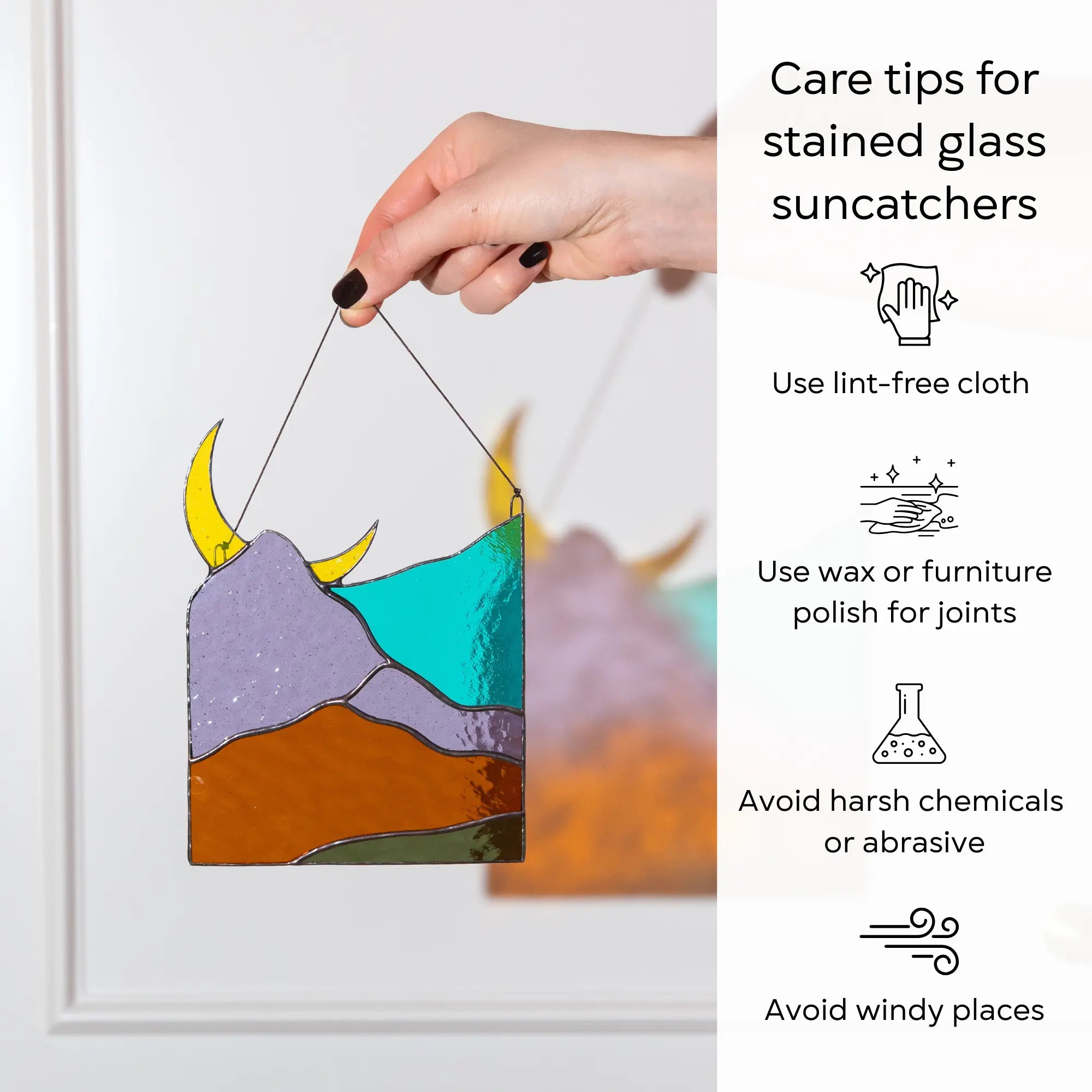 A hand holding a mountain stained glass suncatcher next to cleaning and care instructions: use lint-free cloth, polish joints with car wax or furniture polish, avoid harsh chemicals, and windy places.