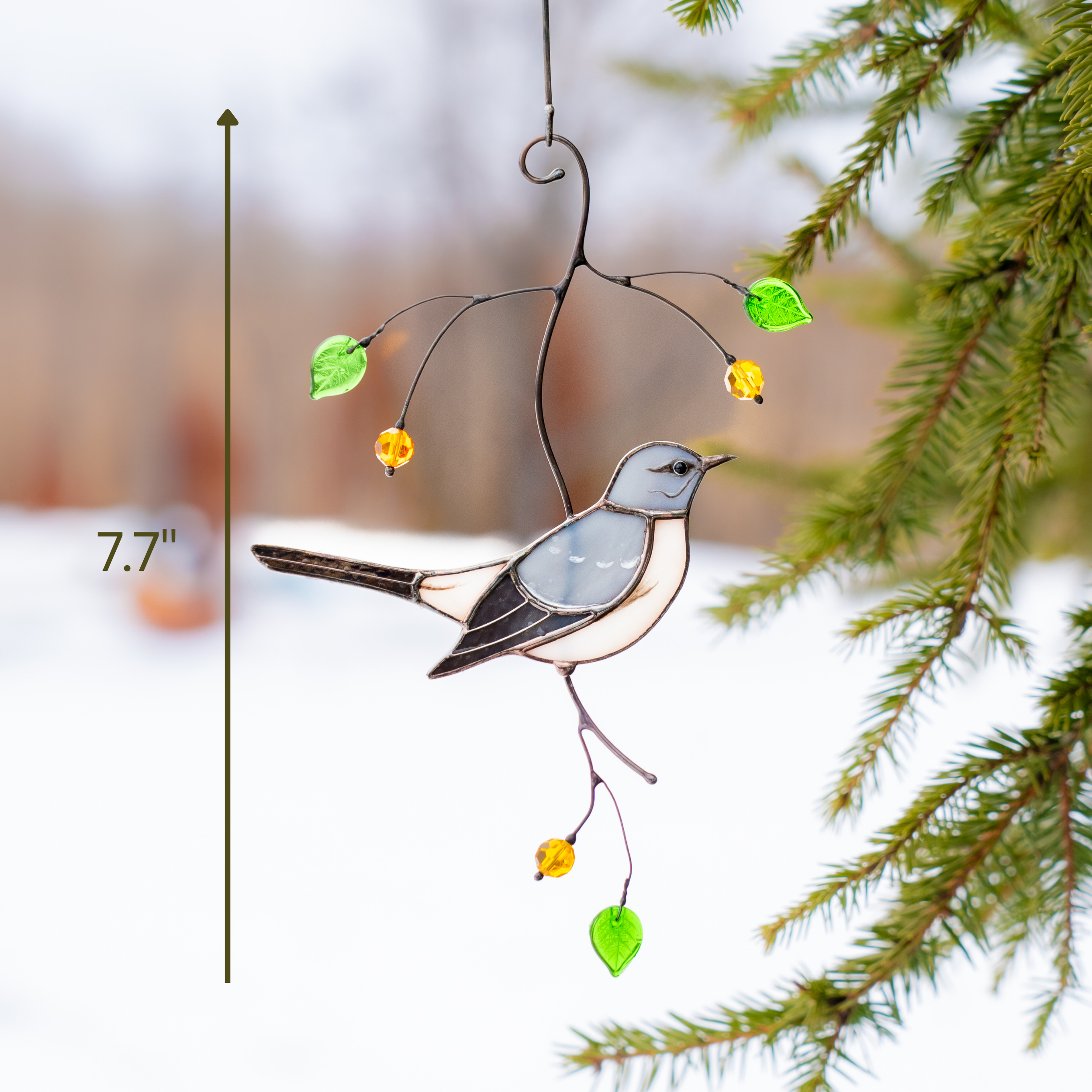 Stained glass mockingbird hanging near pine branches outdoors with snow, showing it's height as 7.7 inches.