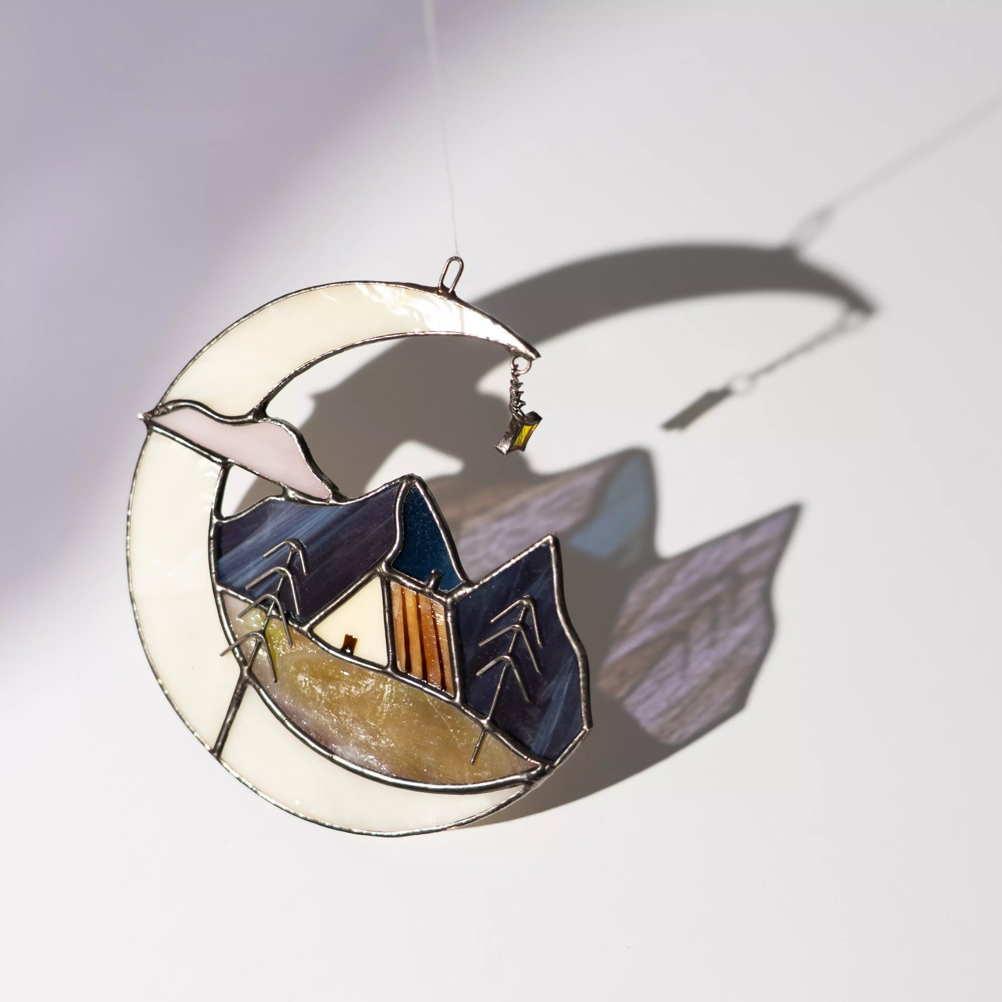 Stained glass moon ornament with a house and trees design casting a colorful shadow on a white surface