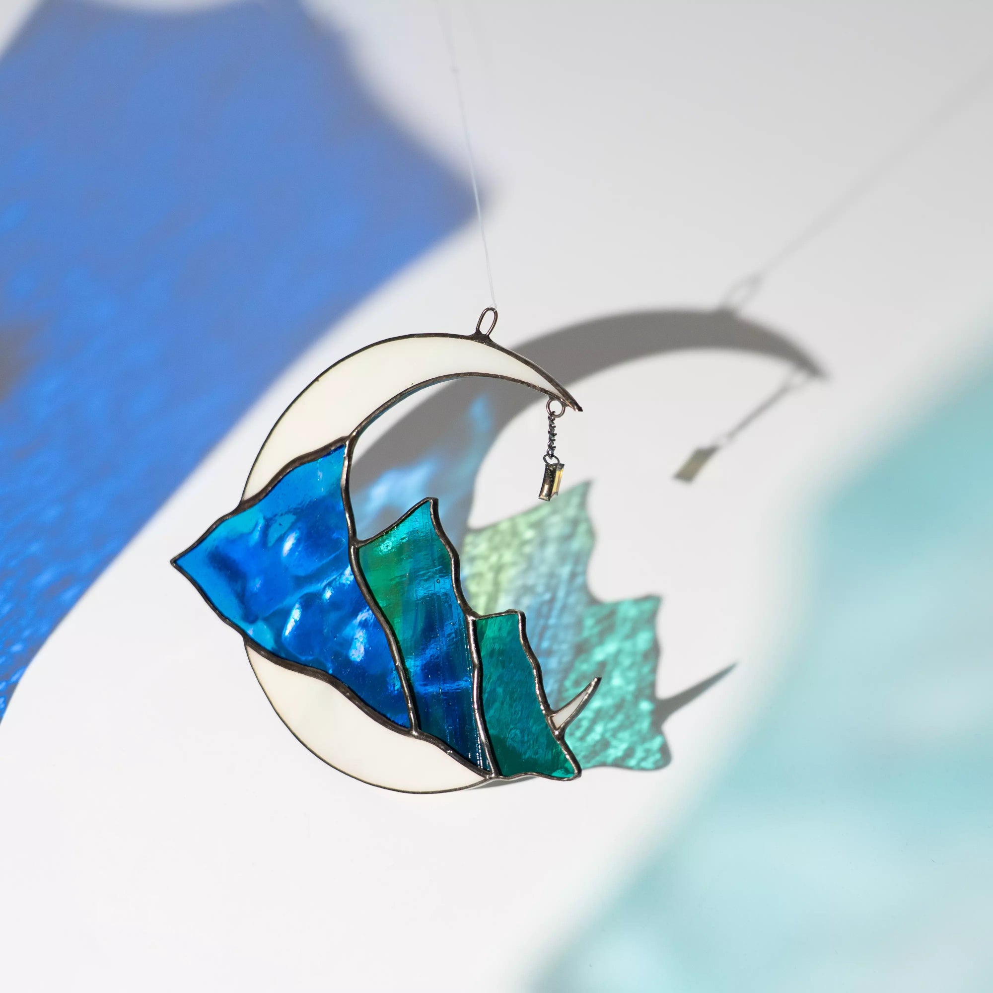 Stained glass celestial suncatcher moon with blue mountains casting a colorful shadow on a white surface