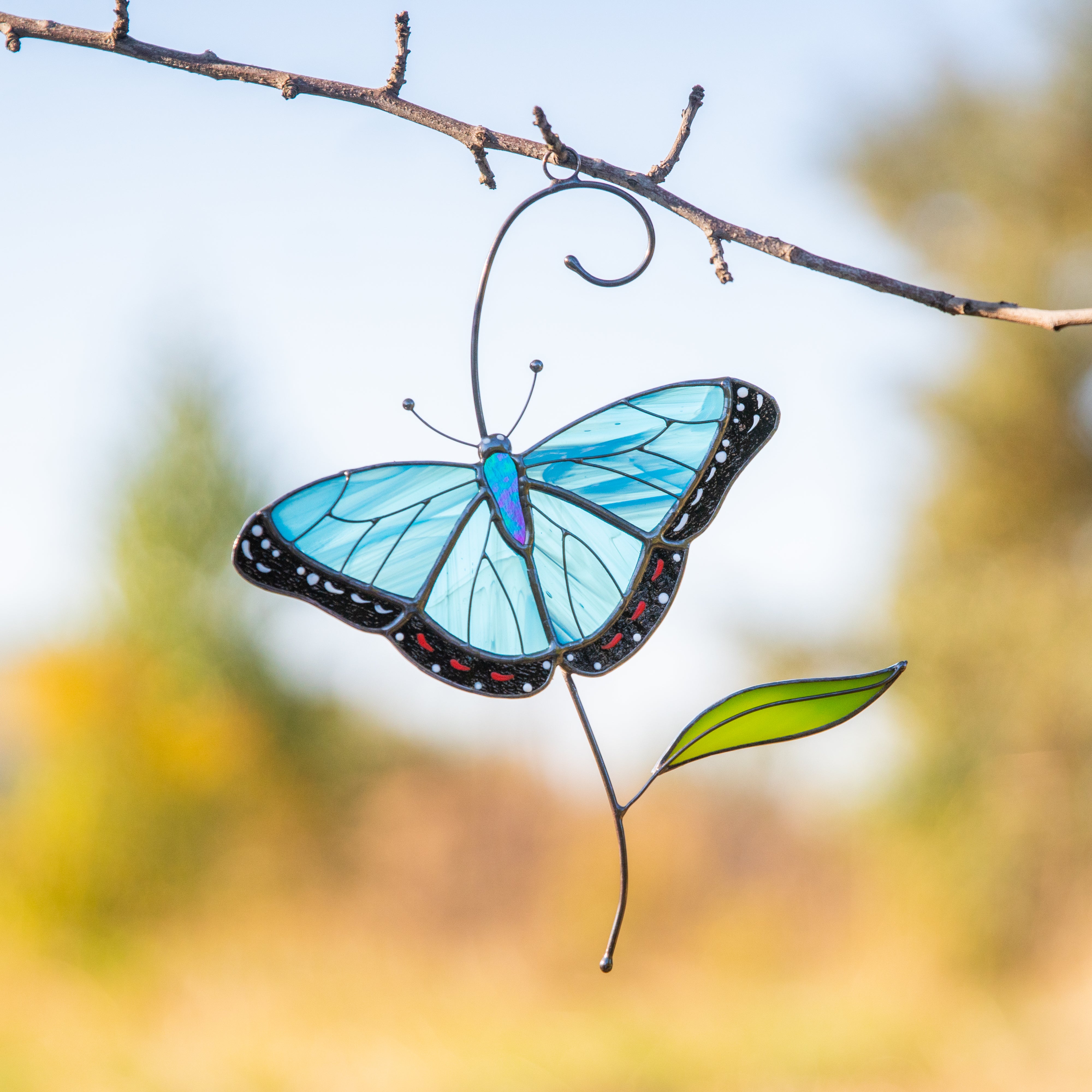 A stained glass morpho butterfly suncatcher with vibrant blue wings and green leaf hanging on the branch against a sunny garden background.