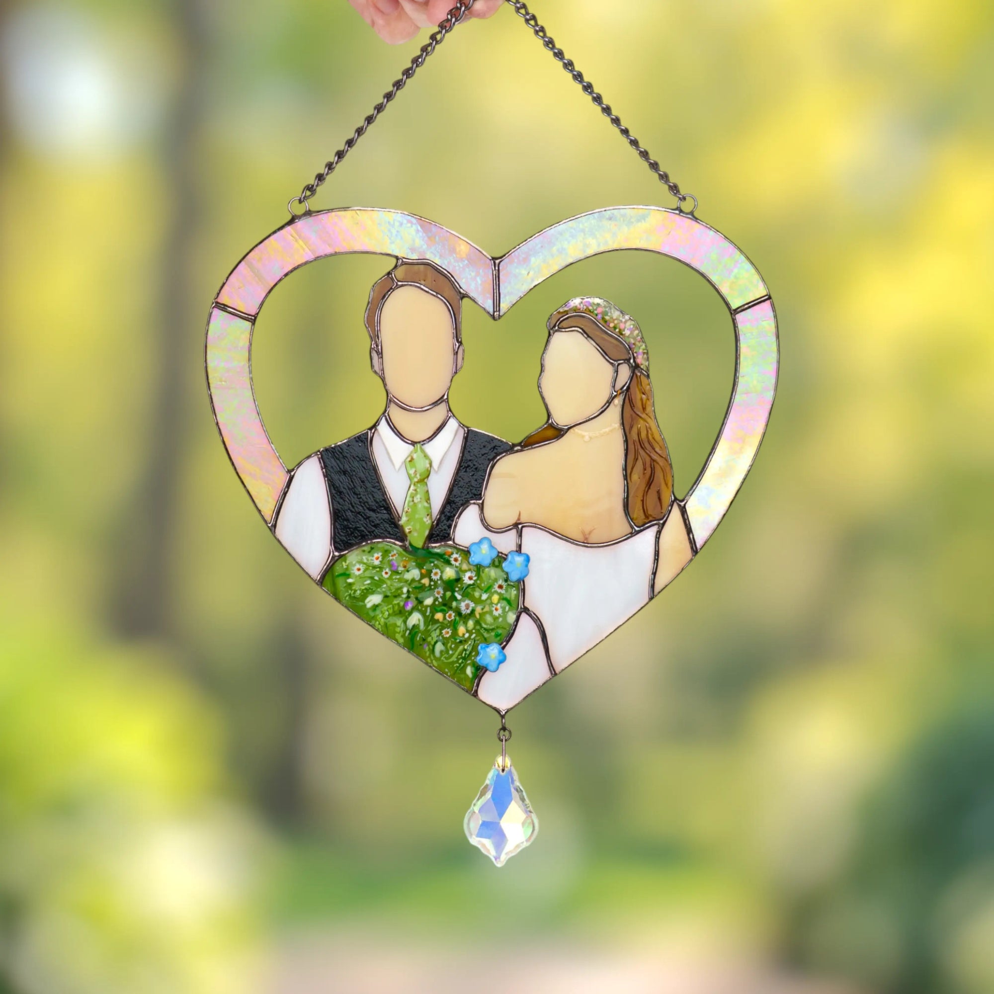 Heart-shaped stained glass ornament with a couple's silhouette and floral details, against a blurred green background.