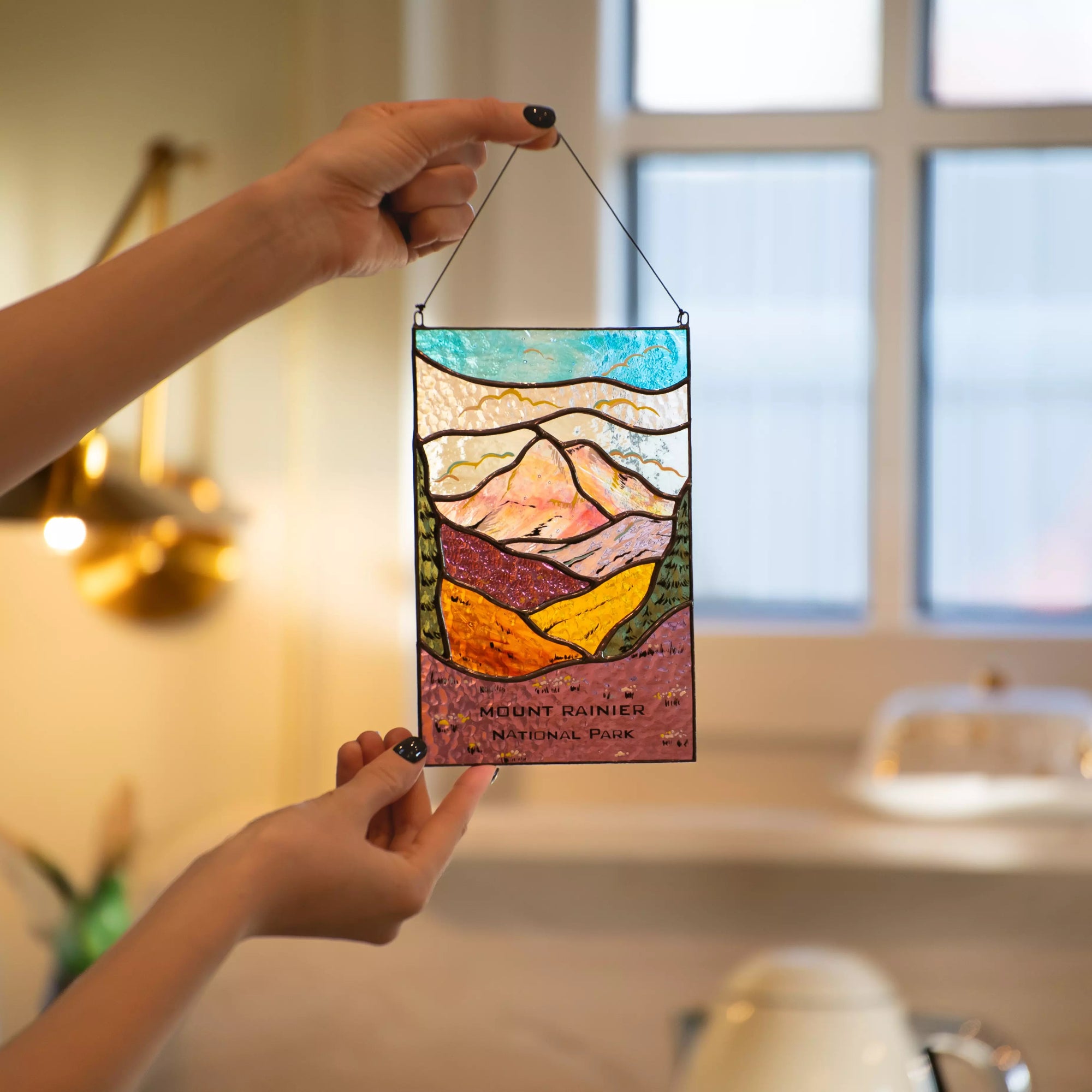 A person holding the Mount Rainier National Park stained glass panel in a cozy interior setting near a window.