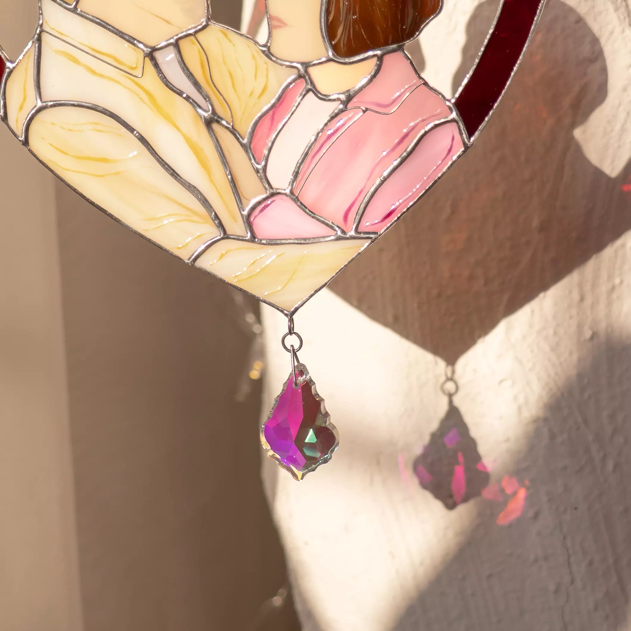 Stained glass window with pink heart and purple teardrop pendant on a textured wall.