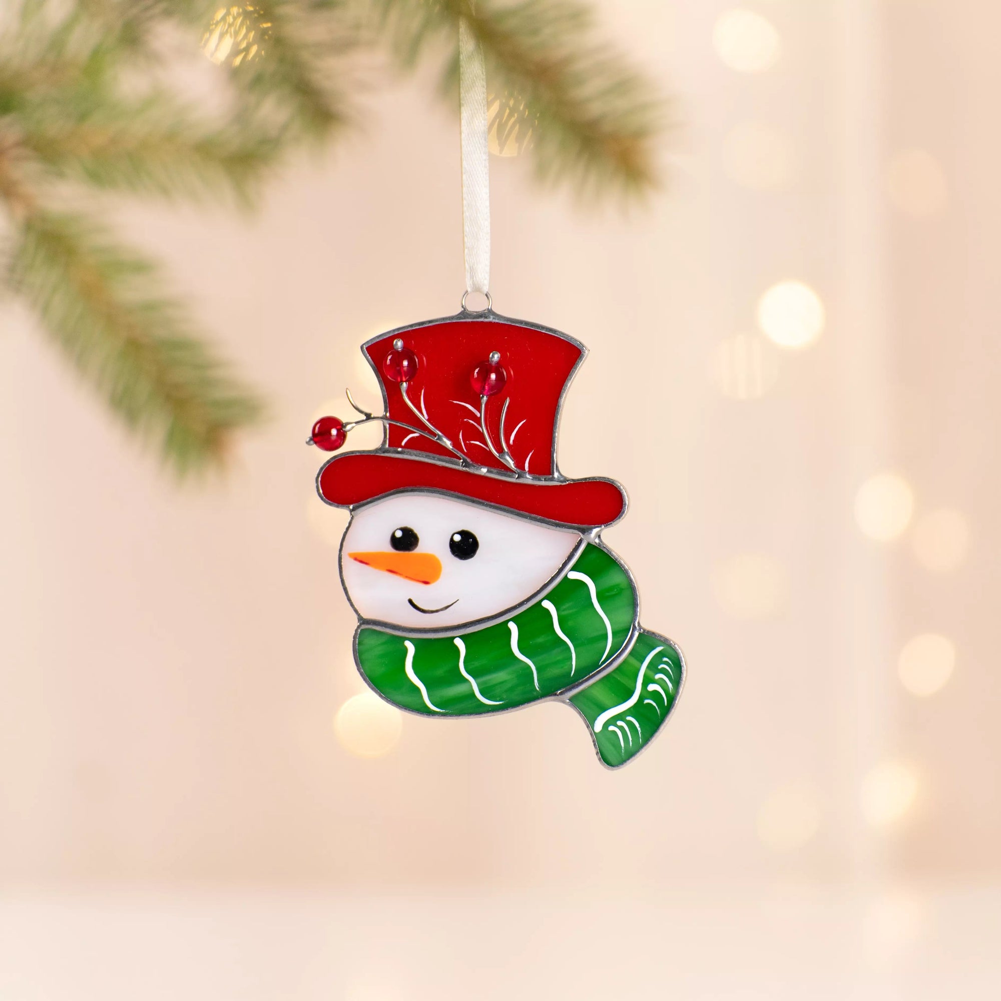 Snowman stained glass suncatcher with red hat and green scarf hanging from a pine branch with glowing lights.