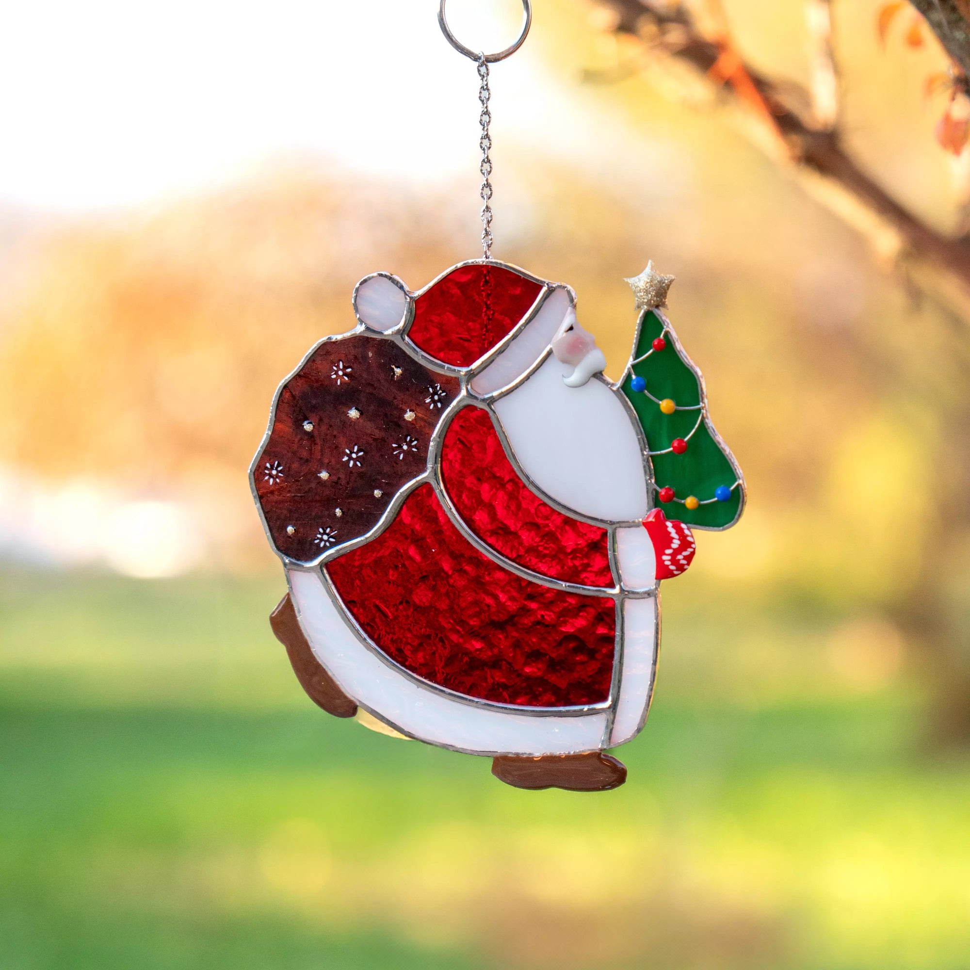 Stained glass ornament of Santa Claus holding a tree against a blurred natural background