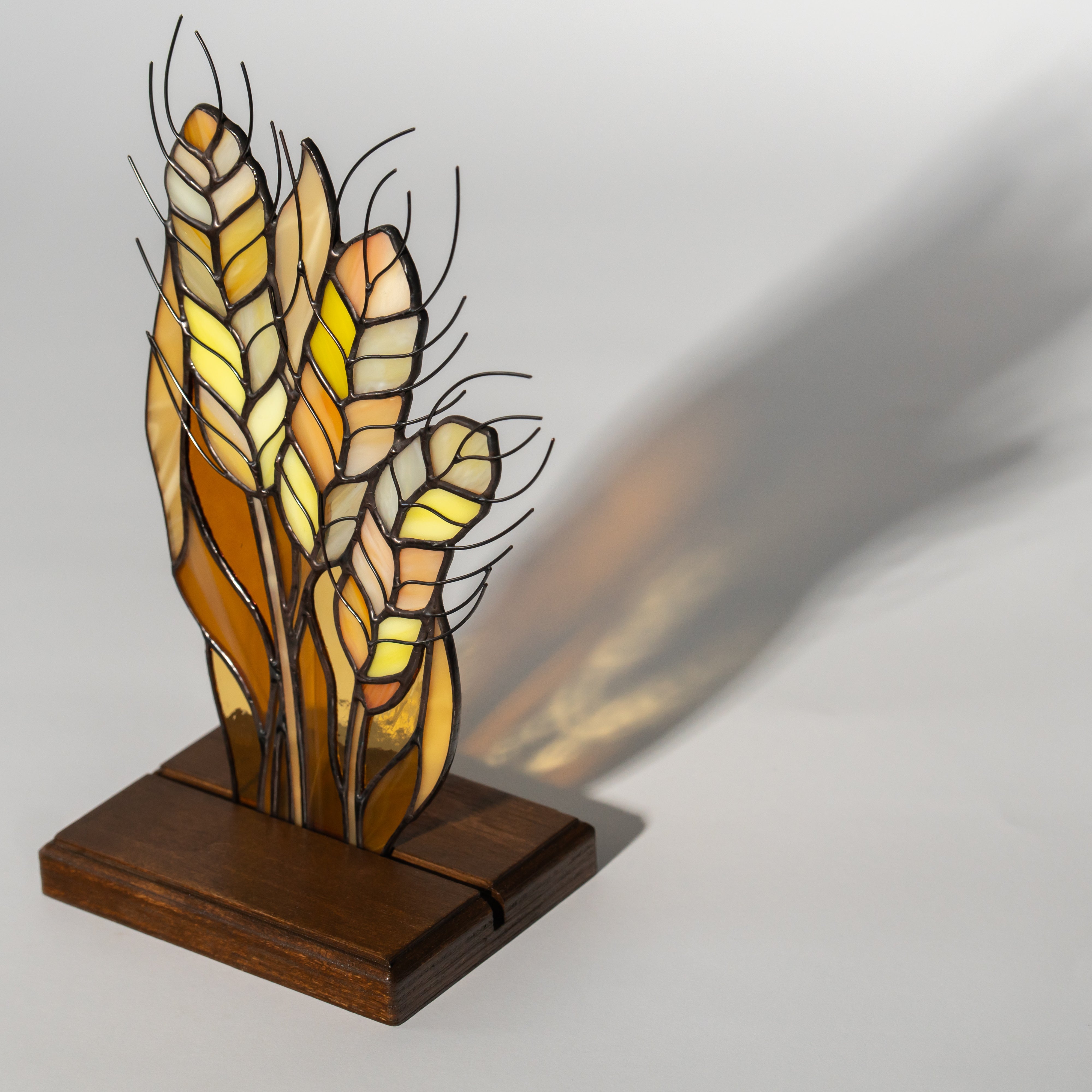 Side-lit stained glass wheat stalks in orange and yellow on a wooden base casting a soft shadow.