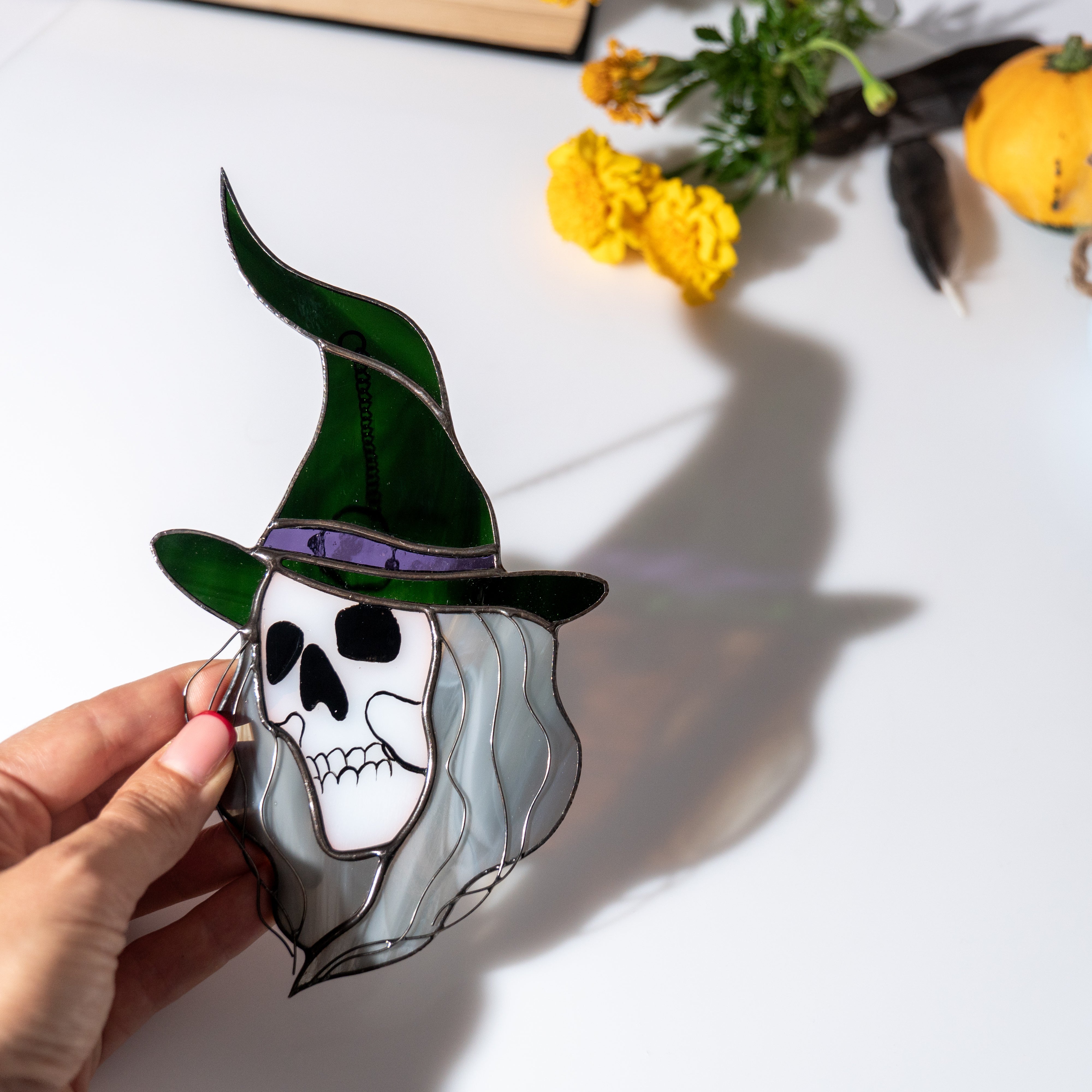 Close-up of a stained glass Halloween witch with a green hat and skull face, casting shadow on a white table with marigolds and pumpkin nearby