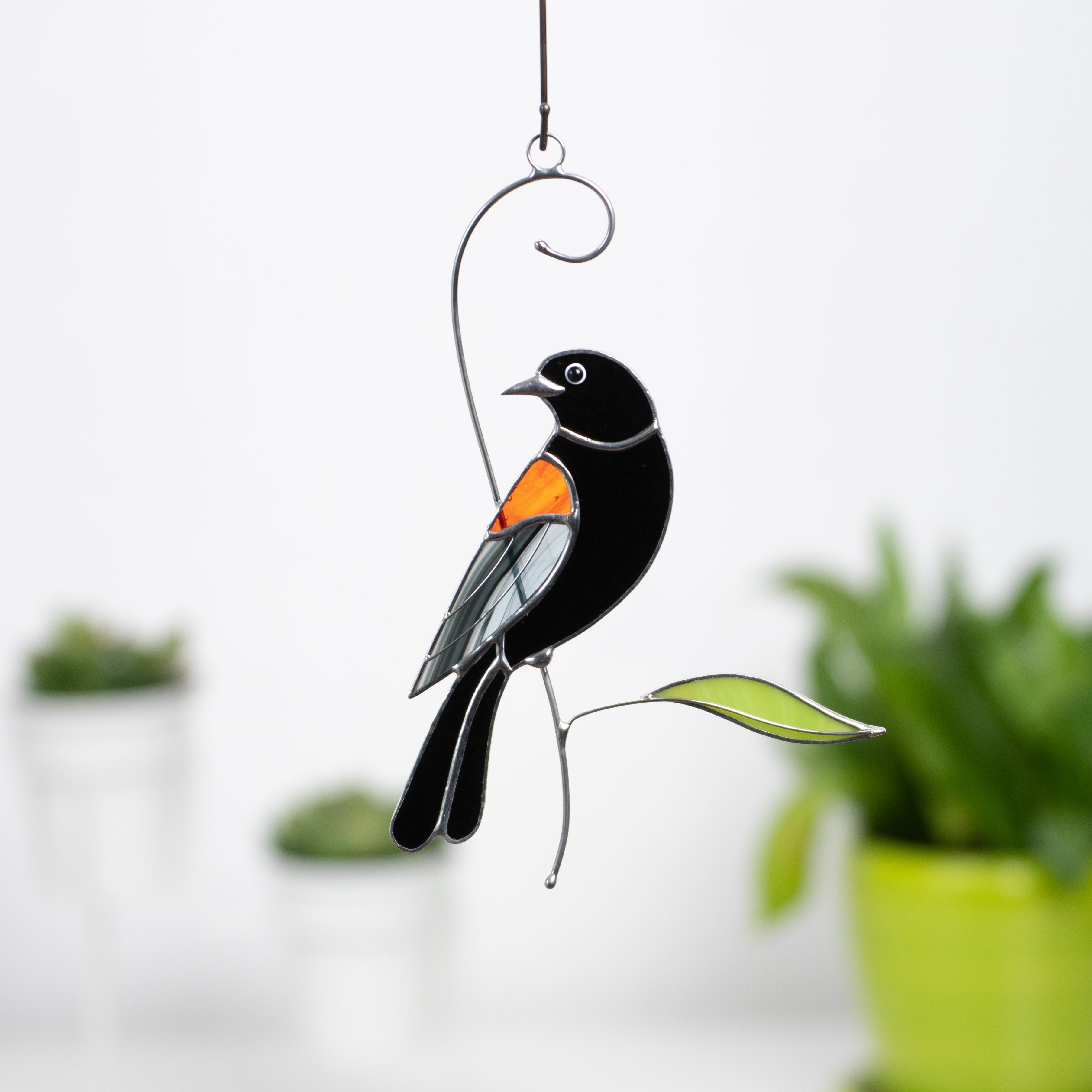 Blackbird suncatcher with vivid orange wing hanging by a green leaf in front of plants.