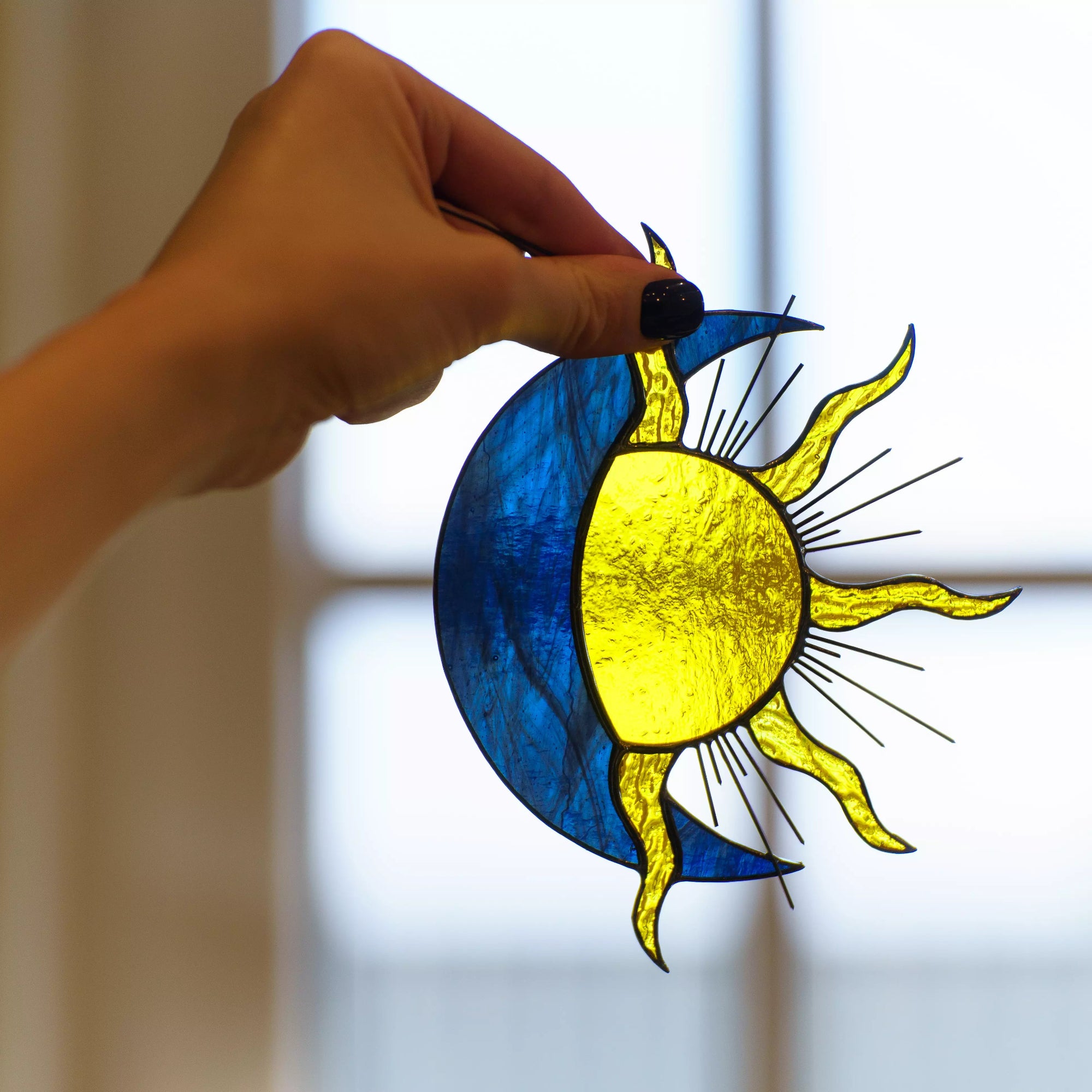 Hand holding a blue and yellow sun and moon stained glass suncatcher against a blurred background