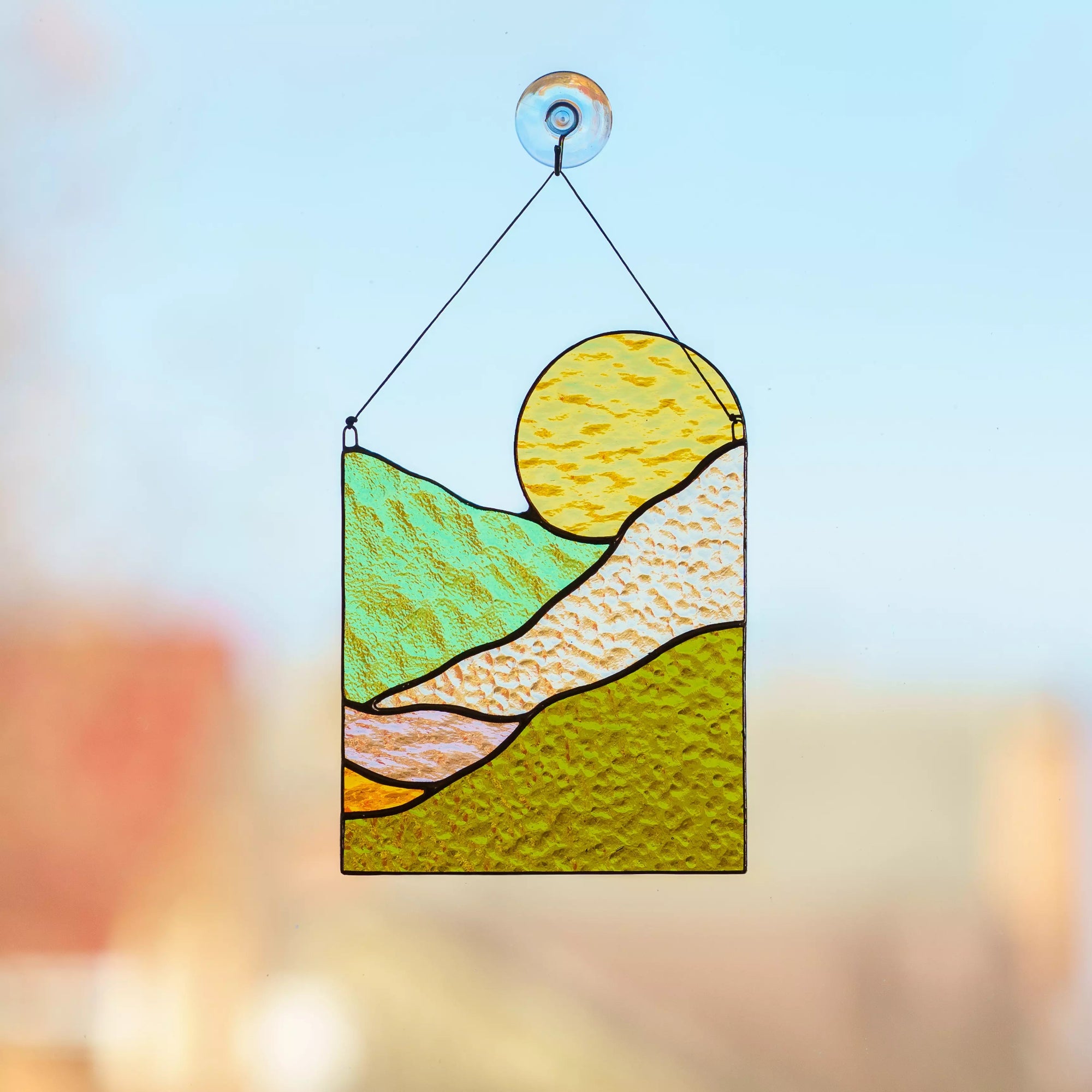 A vibrant stained glass suncatcher with a sun and hills design hanging against a bright sky.