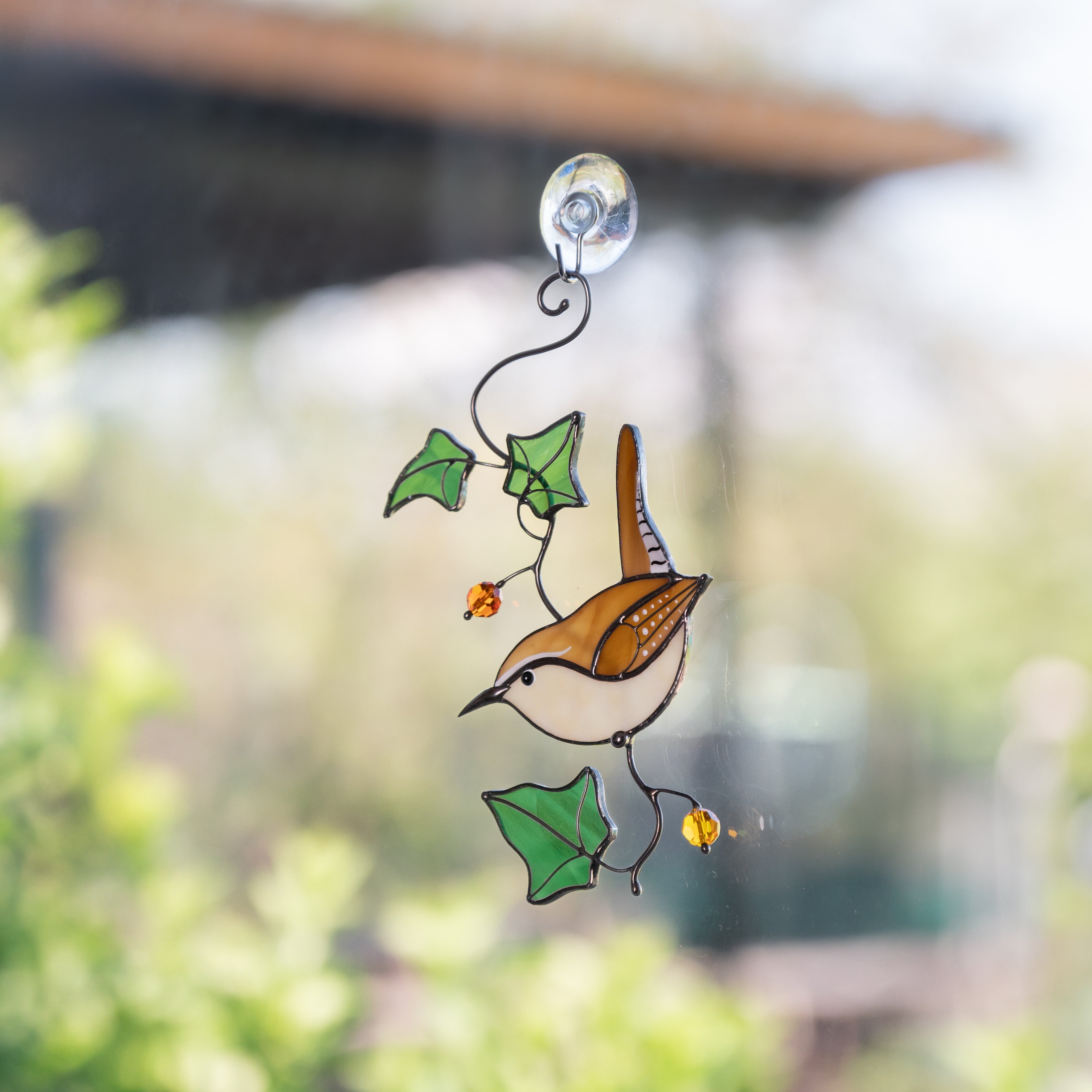 Carolina wren suncatcher hanging on a bright window, highlighted by green leaves and amber beads.