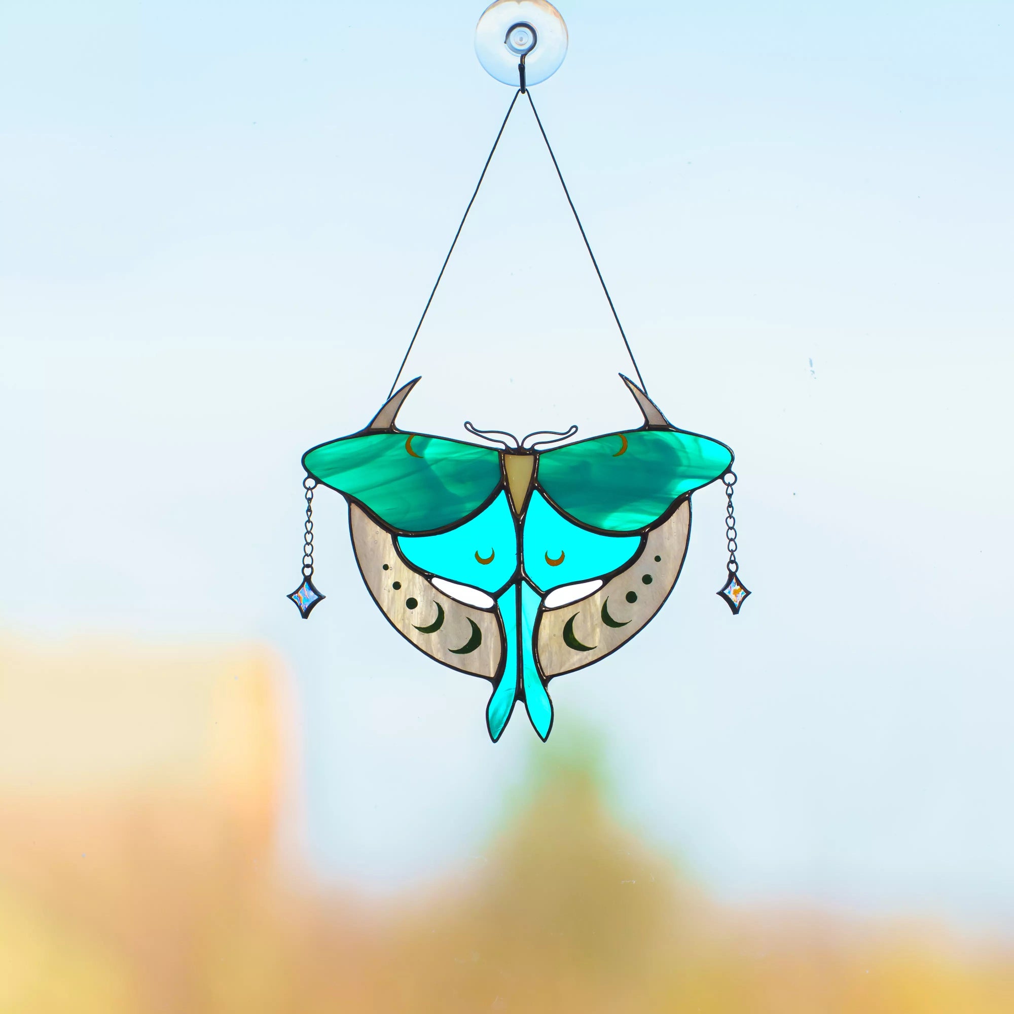 A teal Luna Moth stained glass suncatcher with crescent moon details hanging against a clear blue sky.