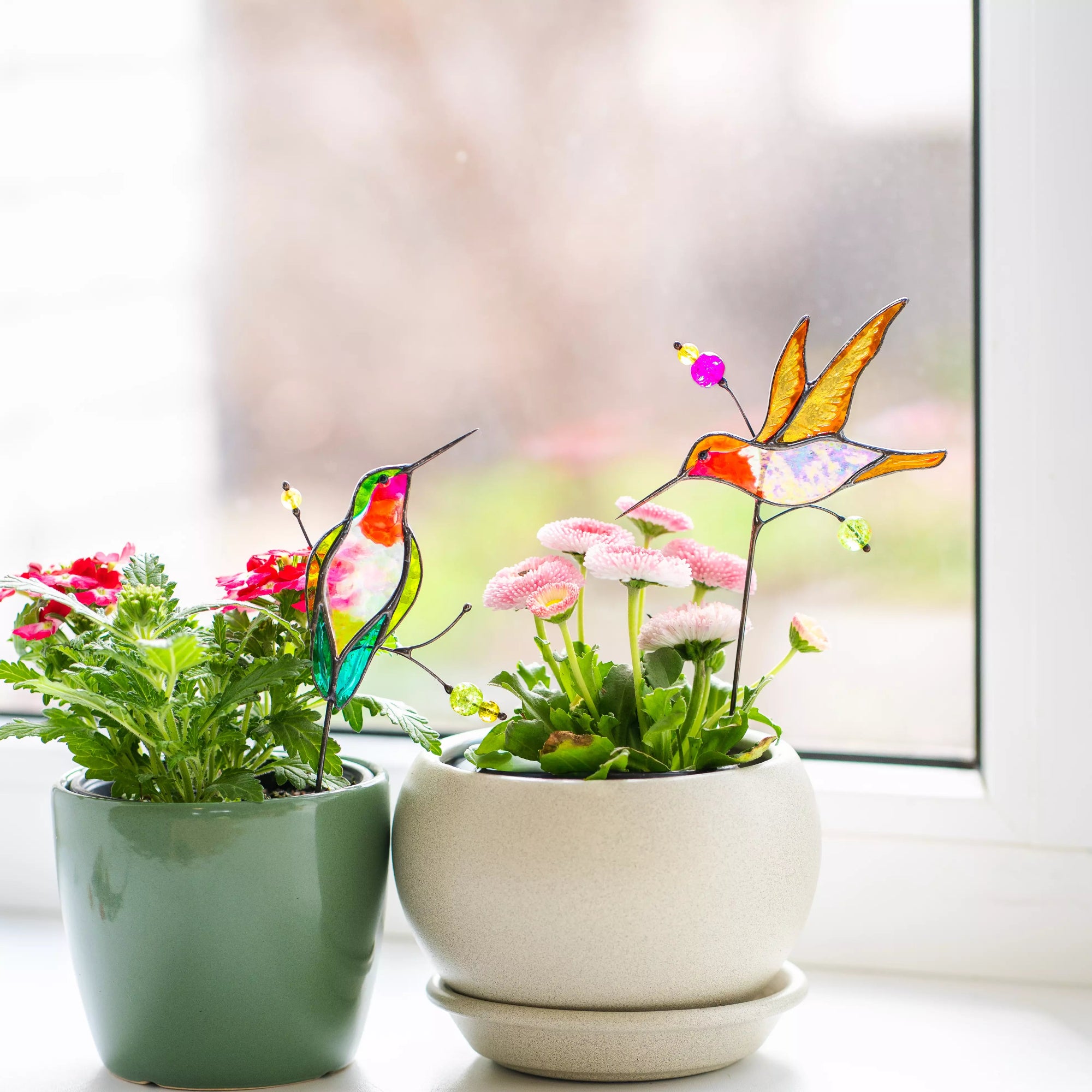 Two potted plants with stained glass hummingbird plant stakes on a windowsill.